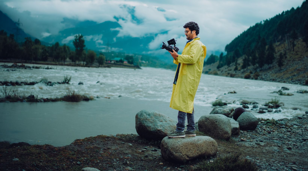 Person in a yellow raincoat standing on rocks by a river, filming a misty mountain landscape.
