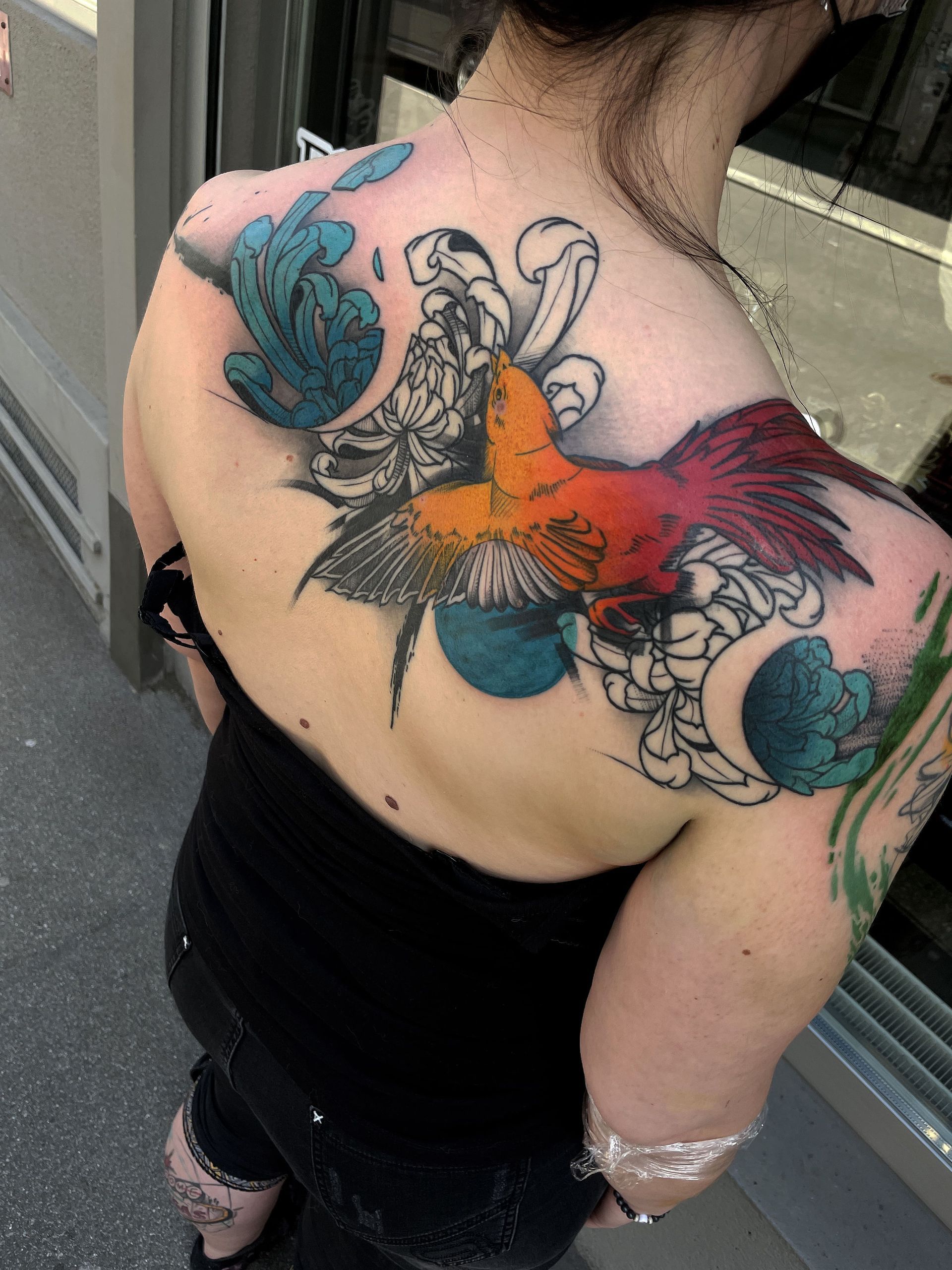 Woman's back tattoo with a colorful bird and abstract floral elements in blue, orange, and red.