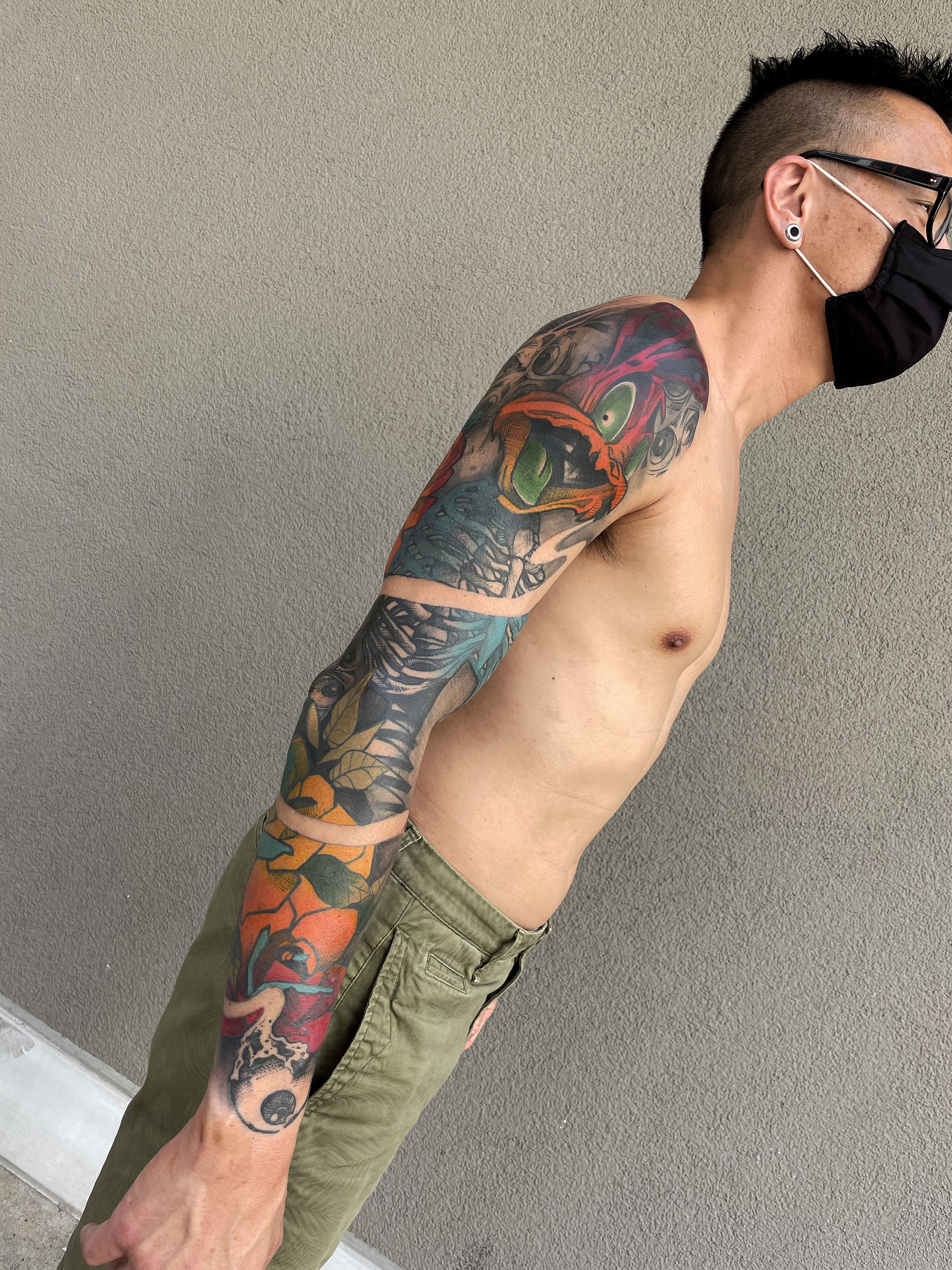 Man with full arm tattoo, wearing mask and green pants, standing against gray wall.