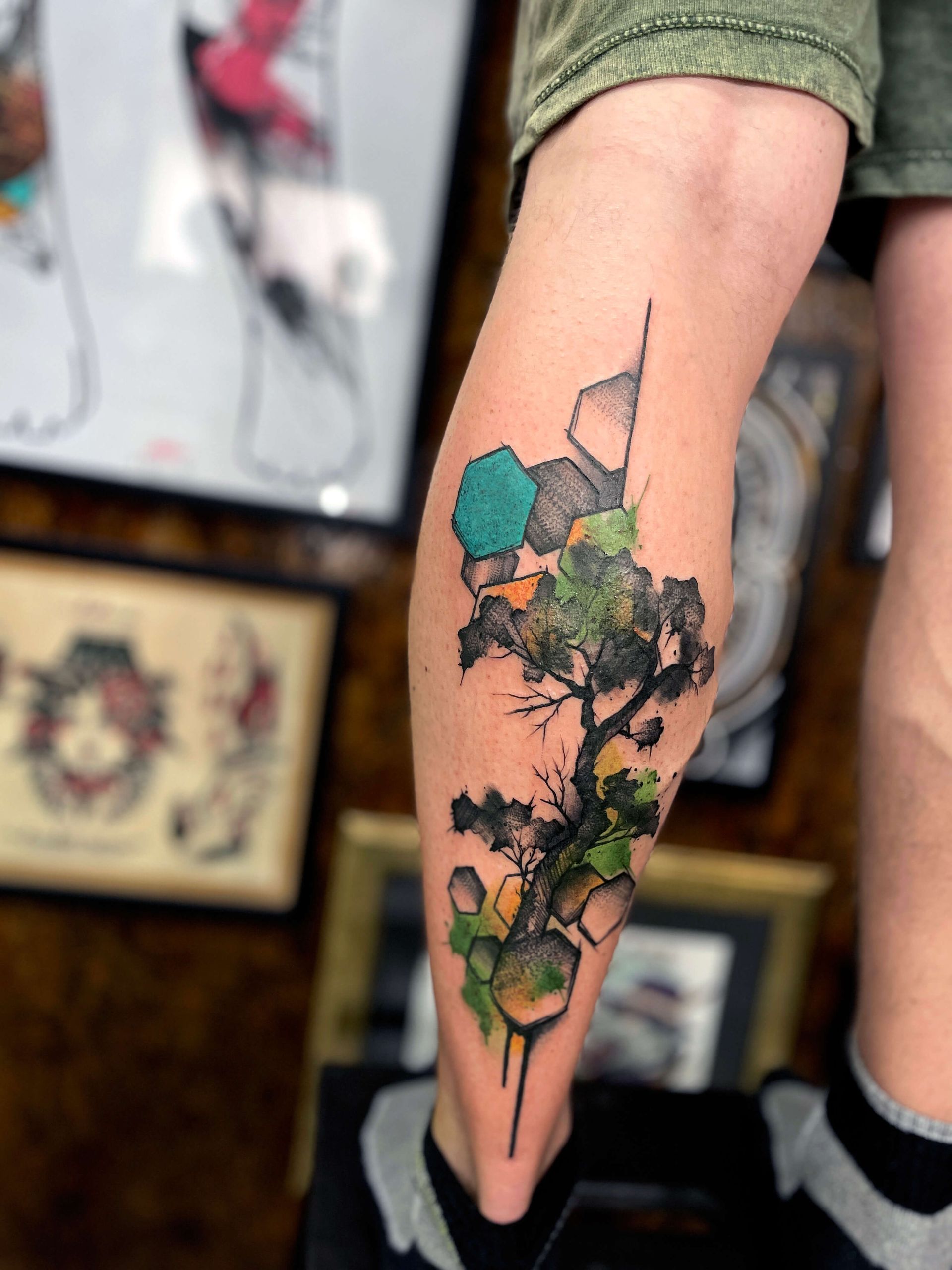 Tätowierung eines Baums mit geometrischen Formen und aquarellartigen Spritzern auf der Wade einer Person.