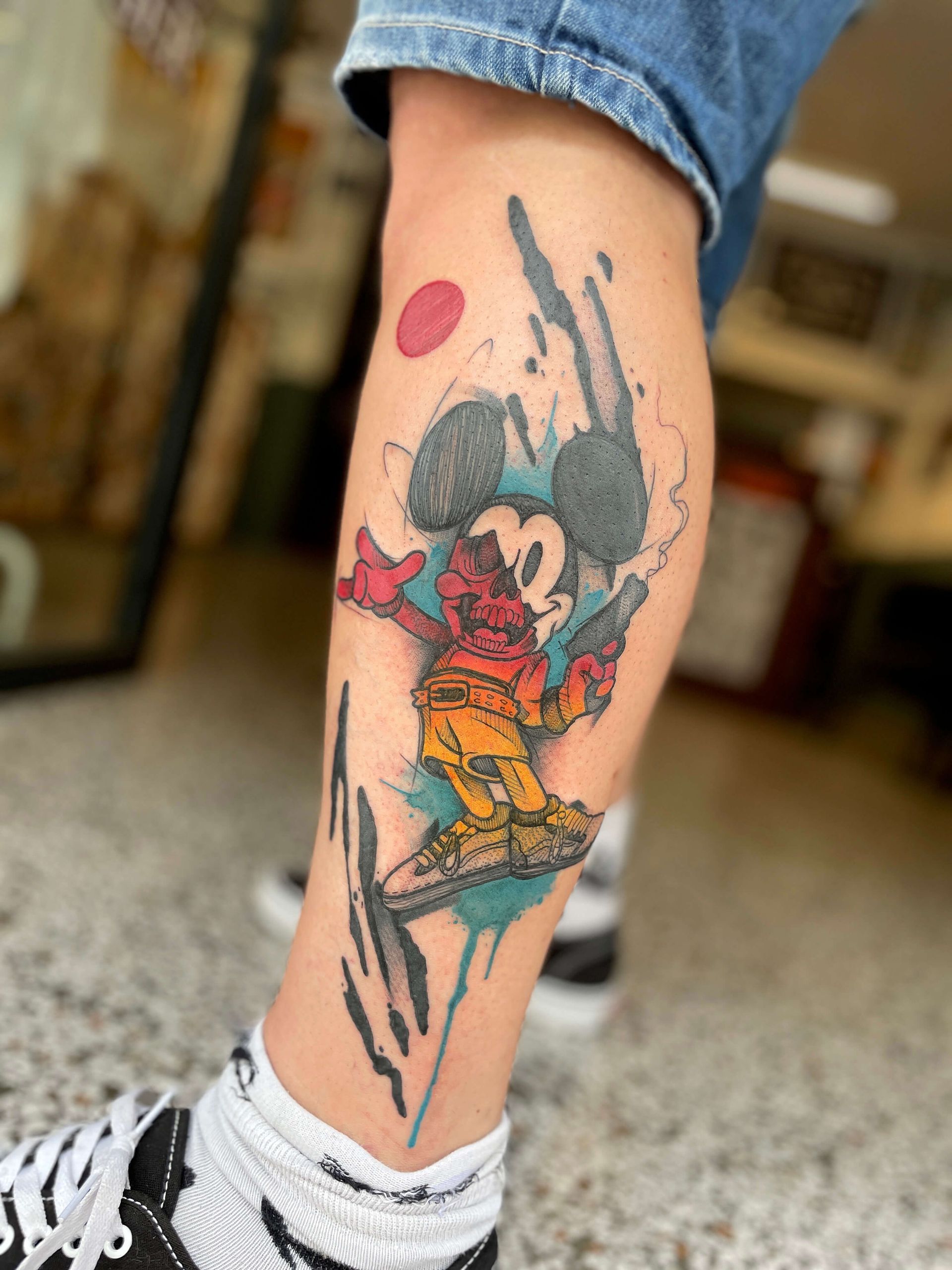 Mickey-Mouse-Tattoo auf einem Bein. Farbenfrohes, cartoonartiges Design mit Spritzeffekt; rot, gelb und blau.