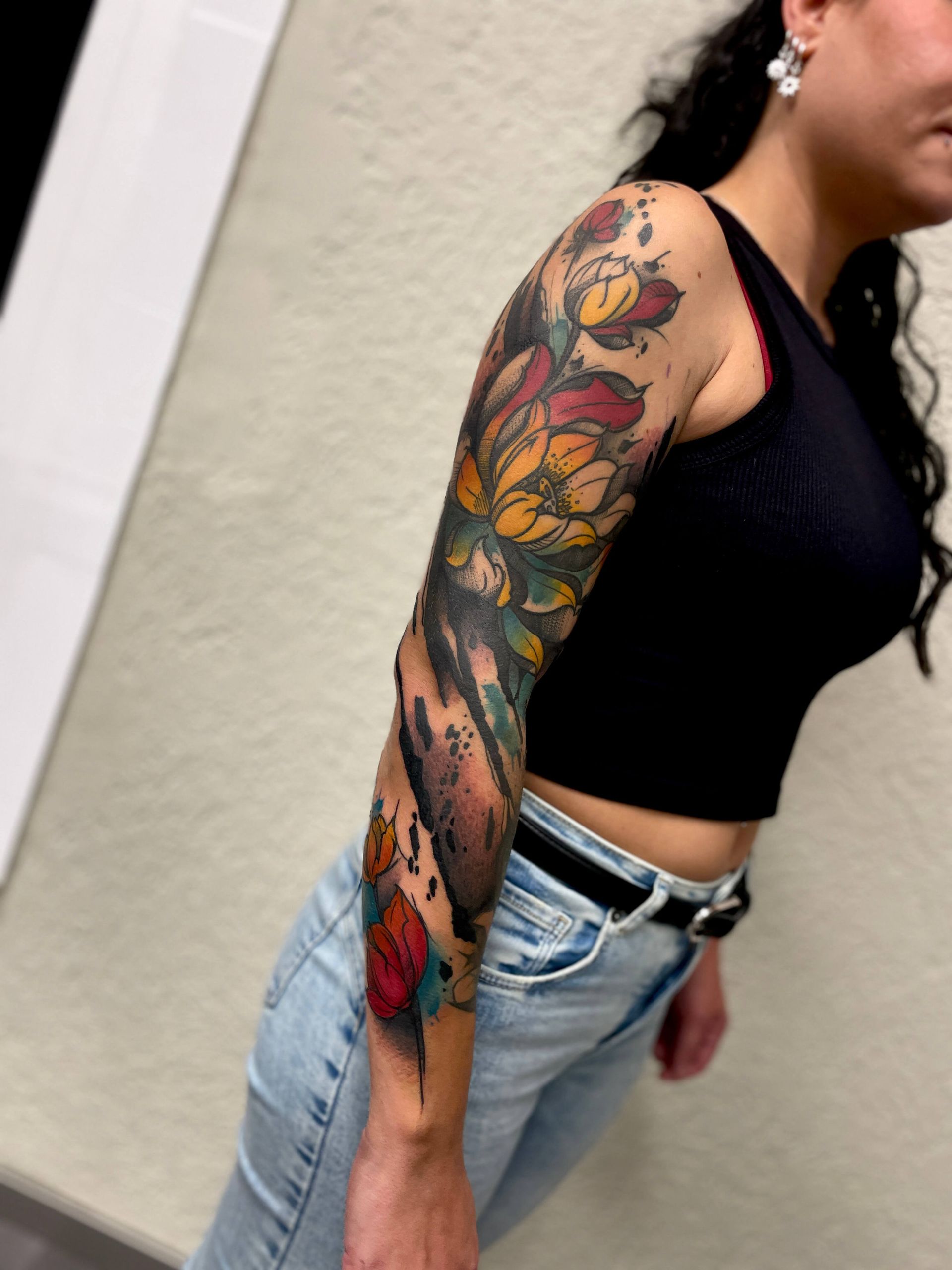 Frau mit einem farbenfrohen Blumen-Tattoo am Ärmel, trägt ein schwarzes bauchfreies Top und Jeans, drinnen.