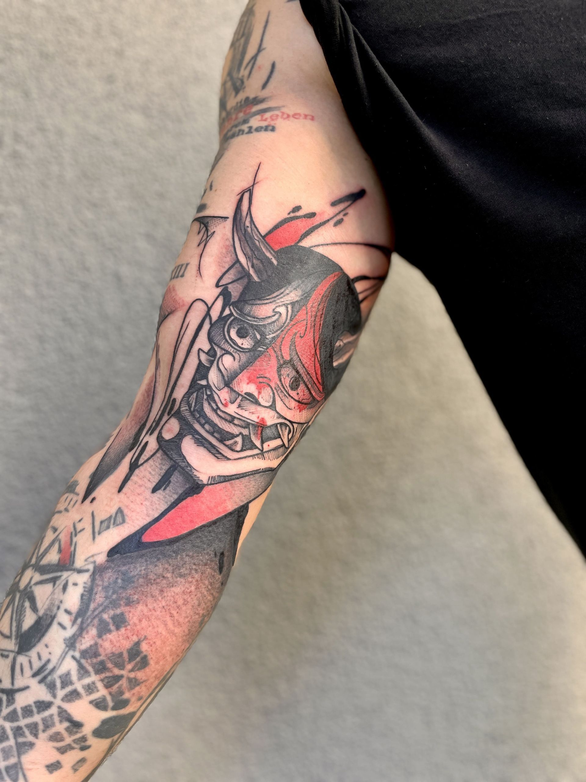 Tattoo of a red and black Japanese demon mask on a person's arm, surrounded by other tattoos.