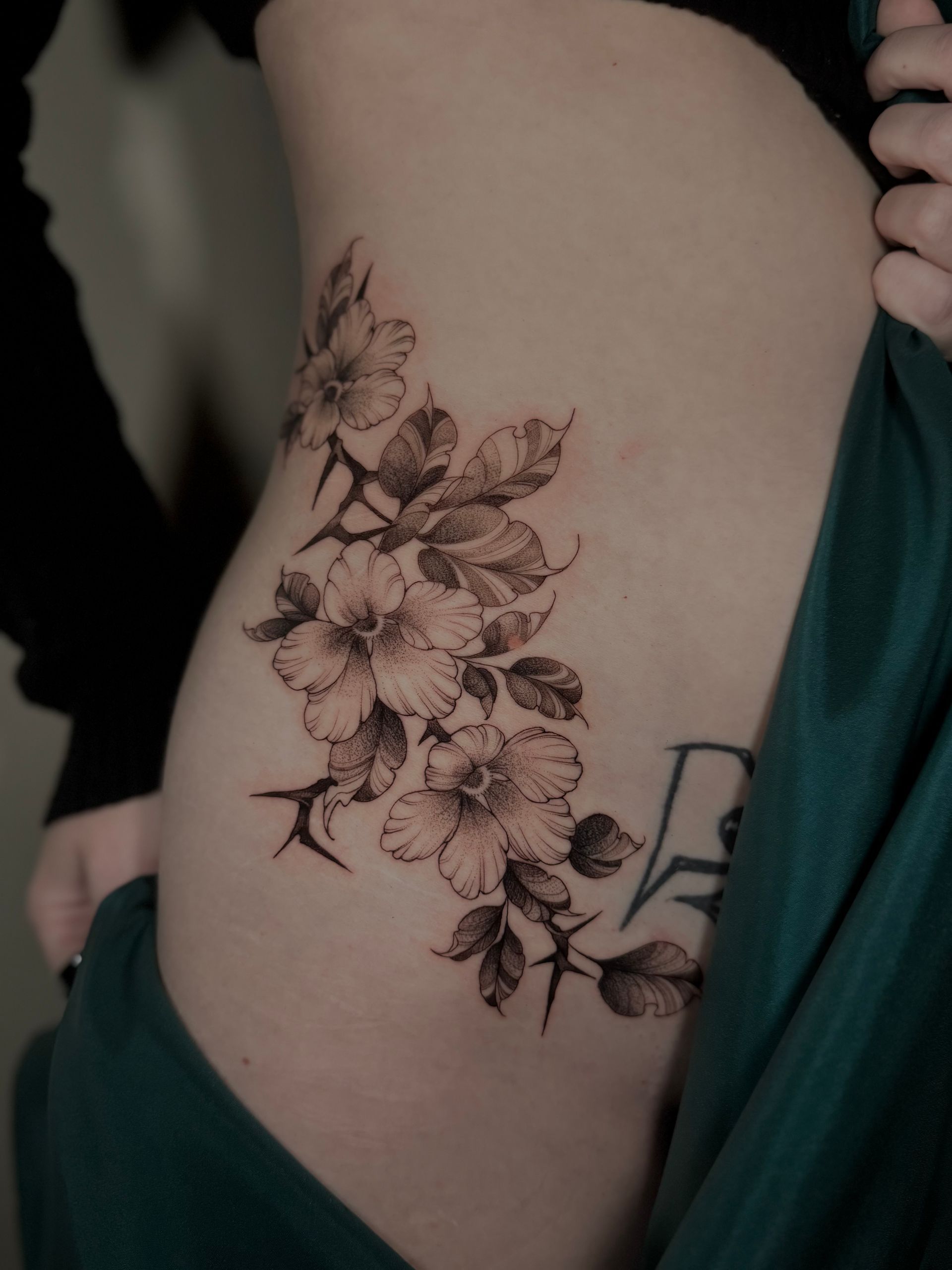 Florales Tattoo an der Seite einer Person, mit Blüten, Blättern und Dornen in Graustufen.