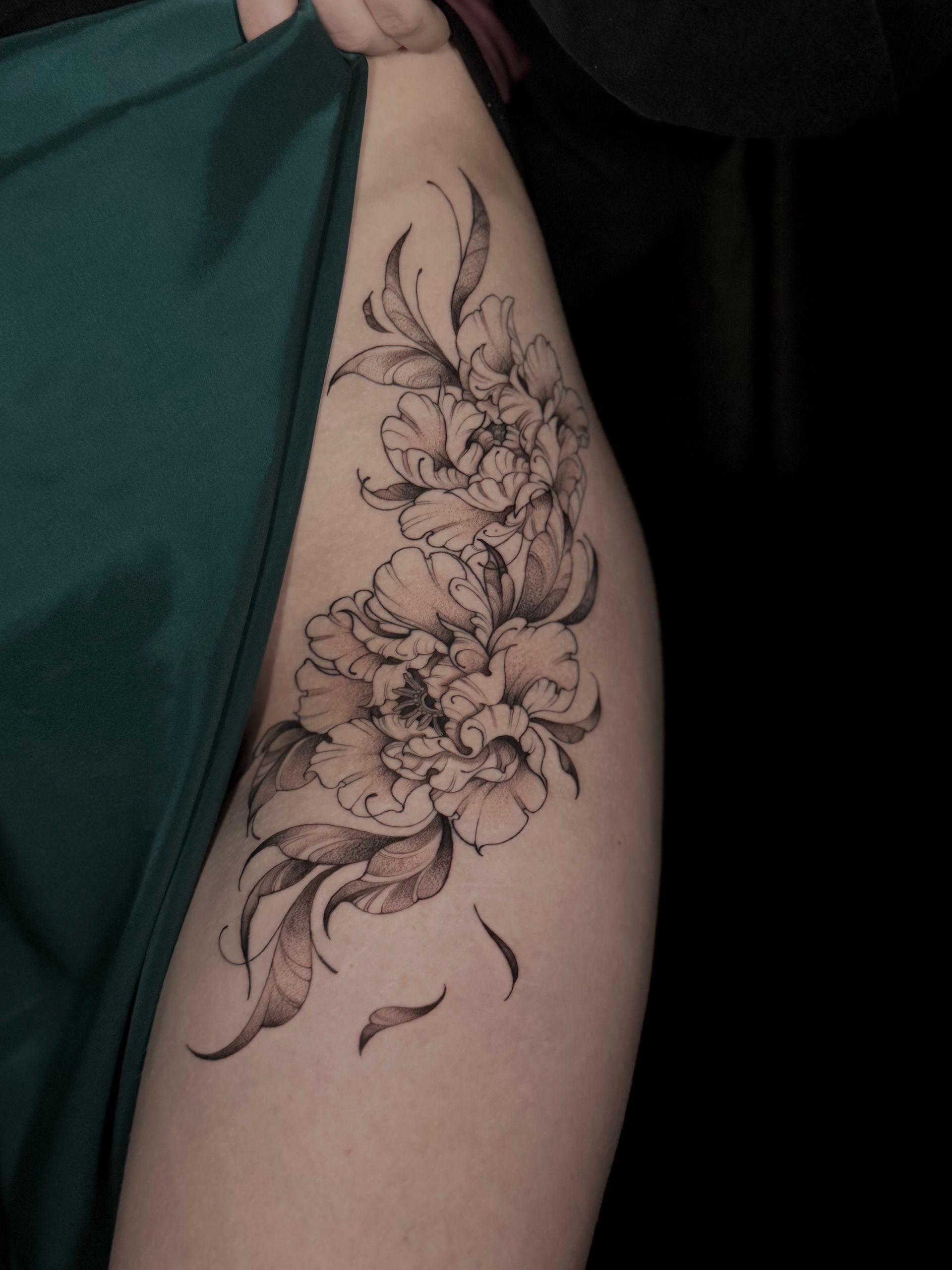 Ein florales Tattoo auf dem Oberschenkel einer Person, mit detaillierten Blüten und Blättern in schwarzer Tinte.