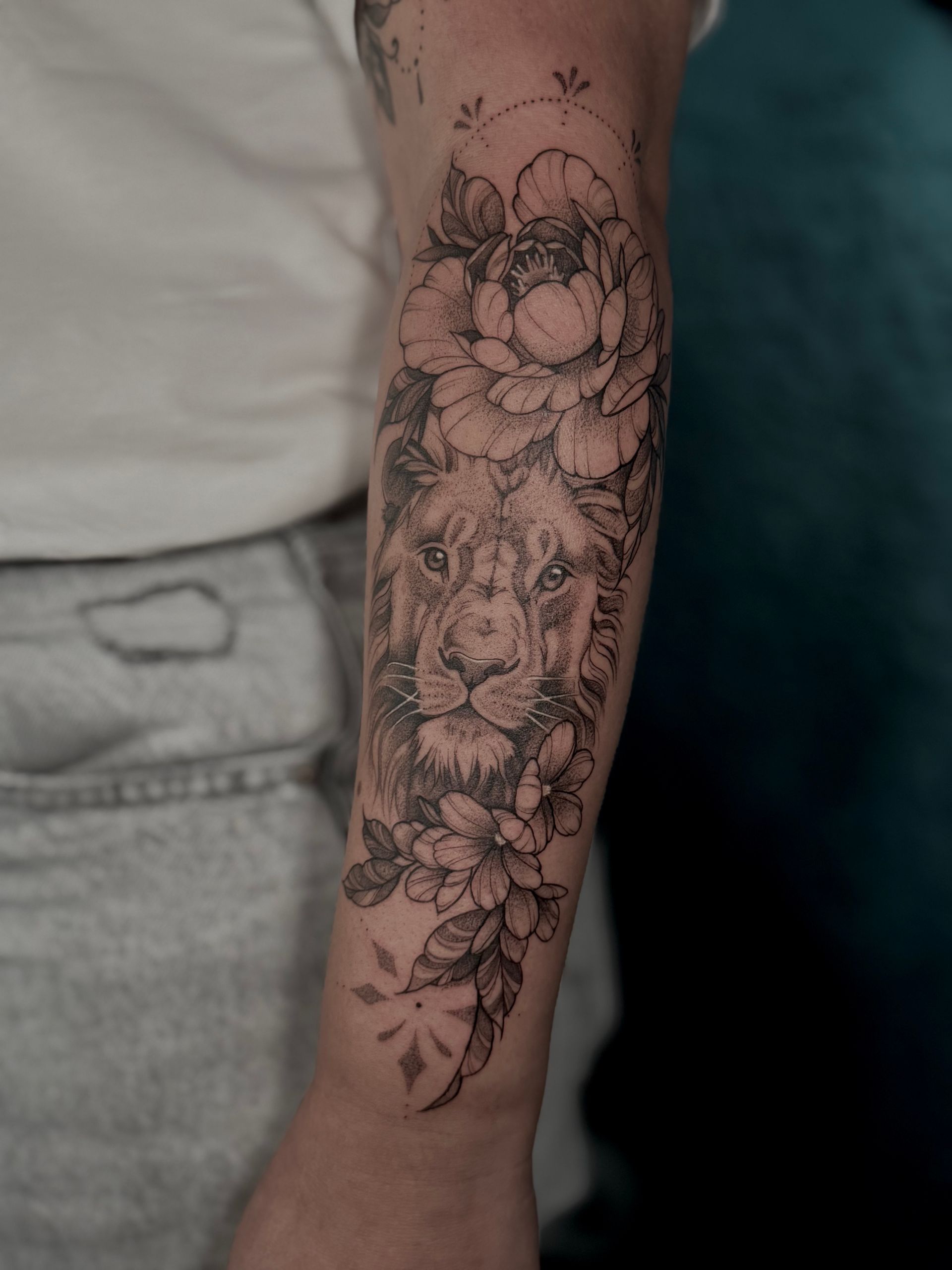 Schwarz-weißes Tattoo eines Löwenkopfes mit großen Blumen auf einem Arm.