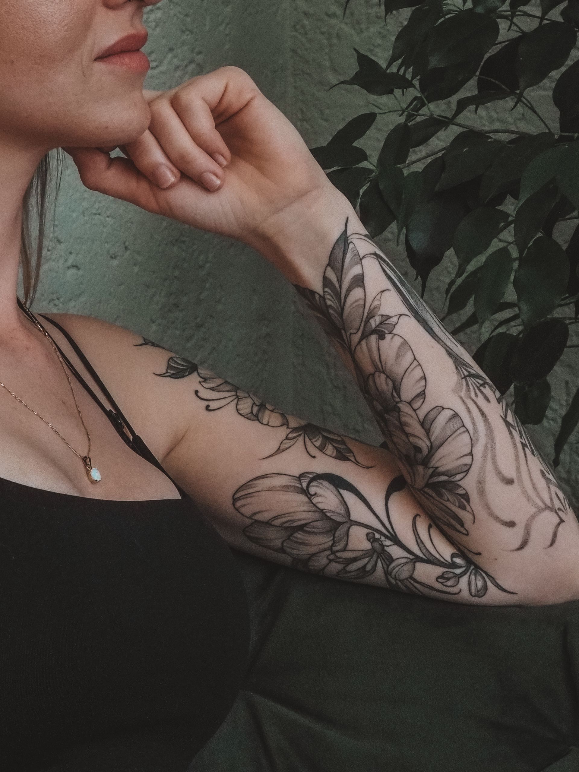 Frau mit floralen Armtätowierungen, das Kinn auf die Hand gestützt. Dunkles Tanktop. Pflanzen im Hintergrund.