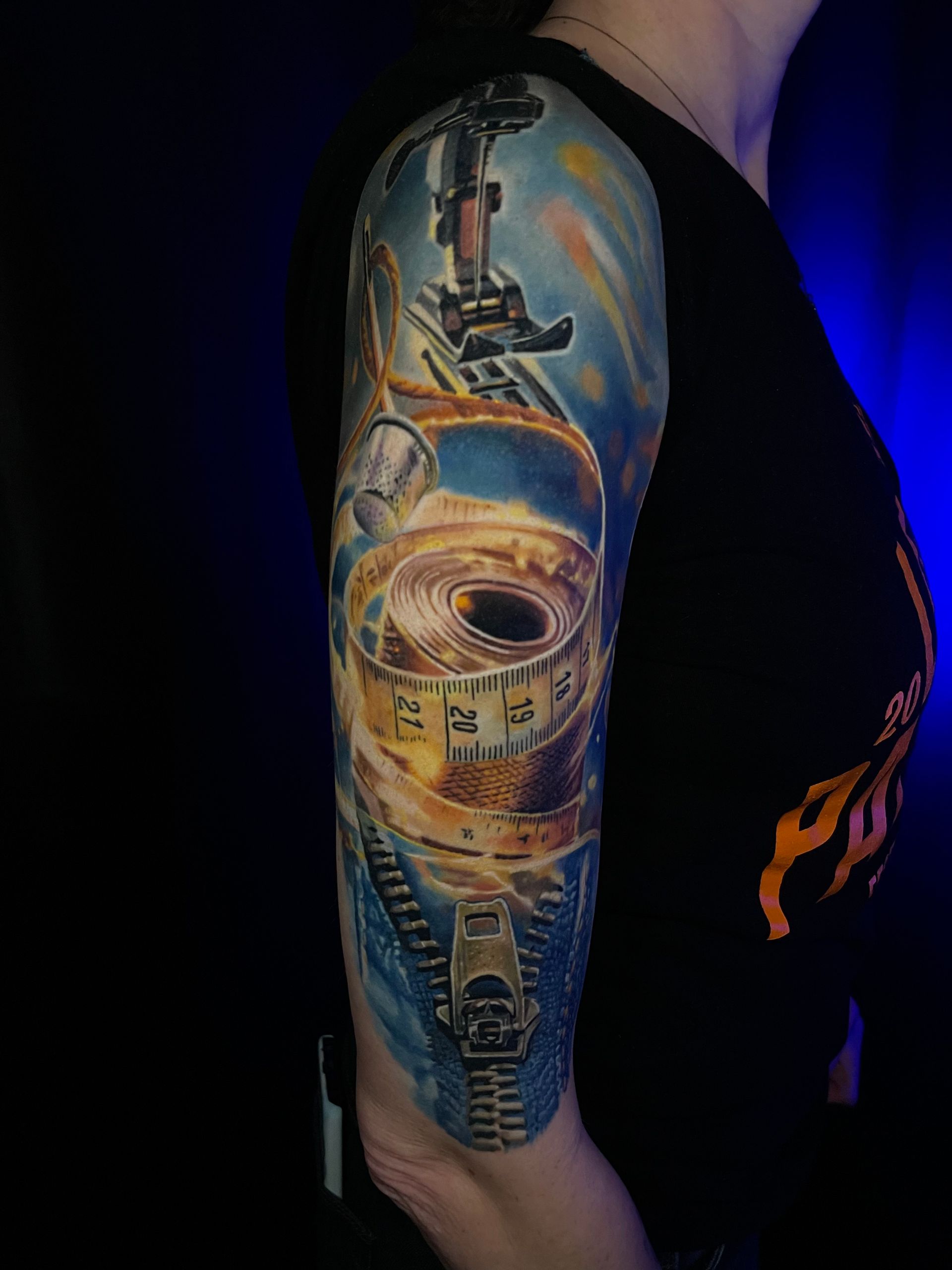 Tätowierung einer Nähmaschine, eines Maßbandes und eines Reißverschlusses auf dem Arm; blaue und goldene Farben auf schwarzem Hintergrund.