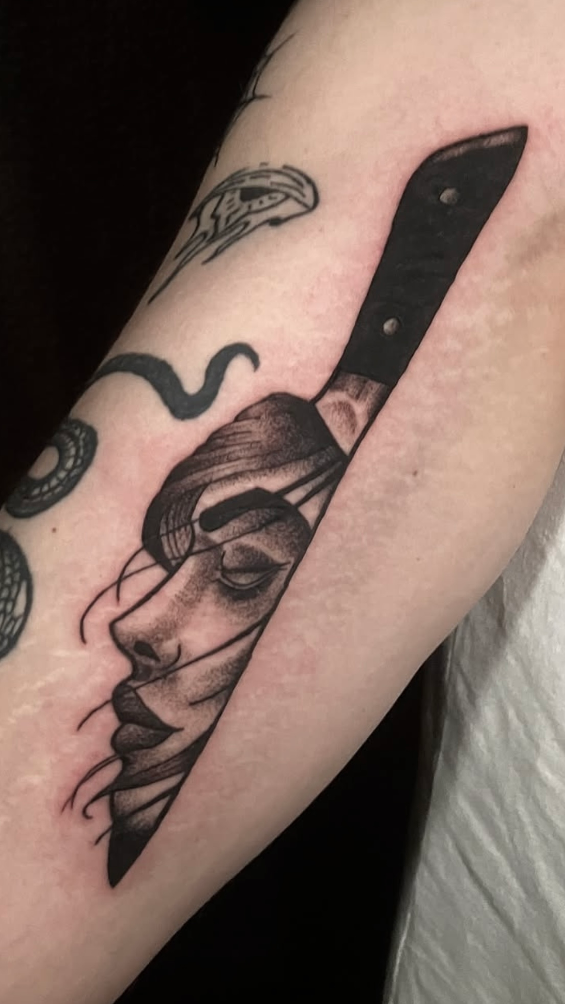 Tattoo of a woman's face sliced by a knife. Black and white with details in shading on the forearm.