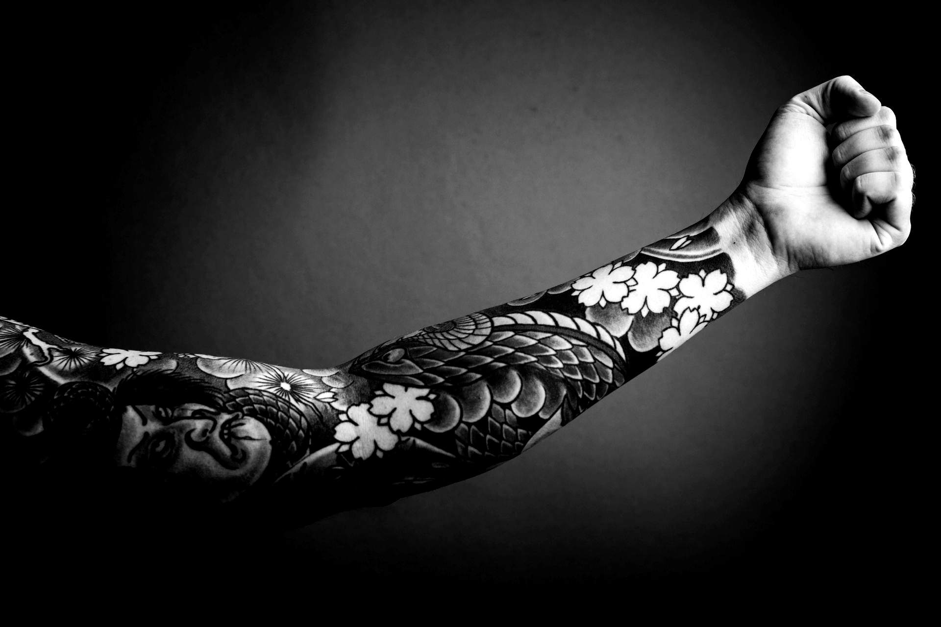 Black and white photo of a tattooed arm, raised in a fist, against a dark background.