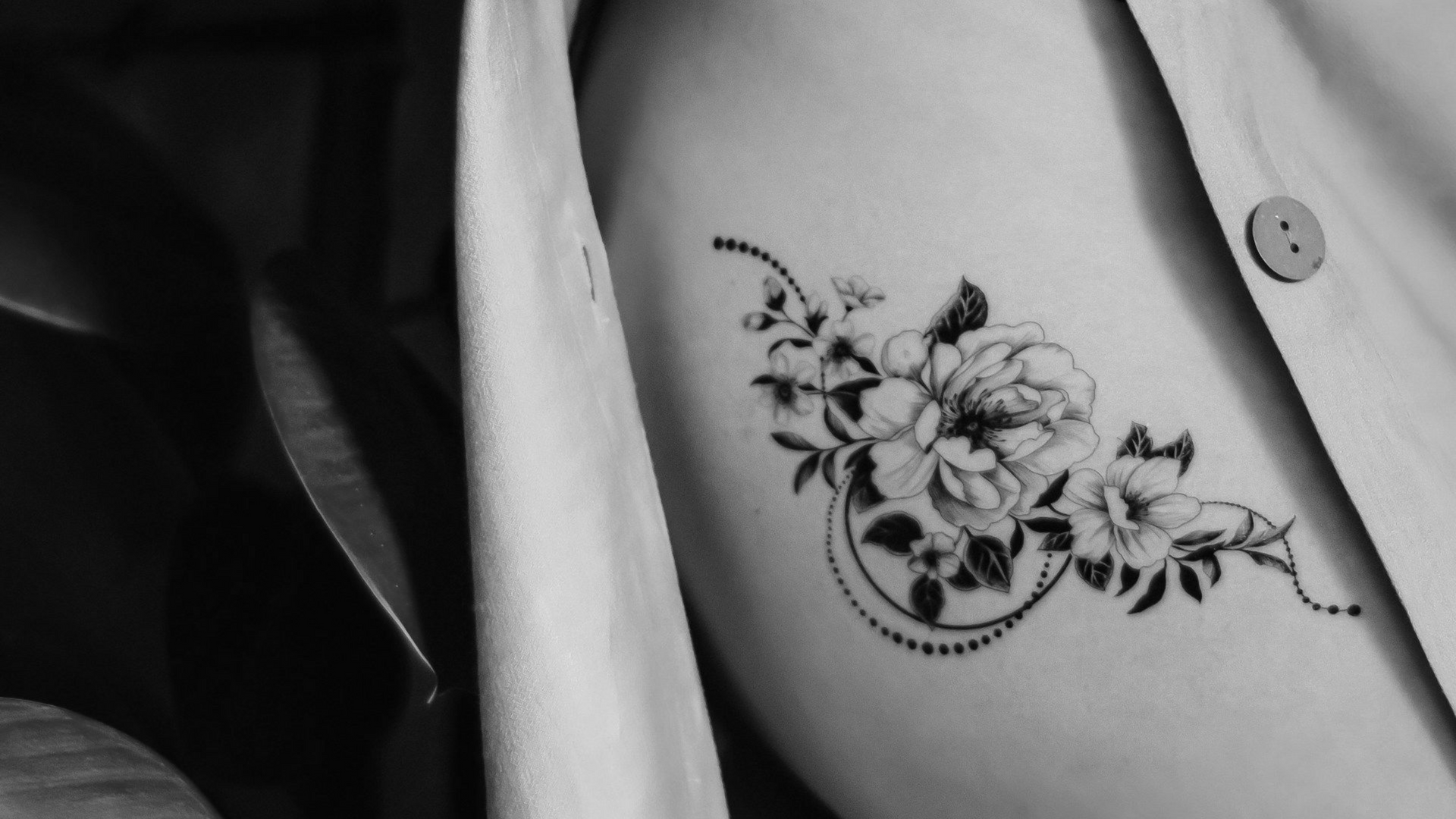 Schwarz-weißes Blumentattoo auf dem Arm einer Person, neben einem Button-Down-Hemd.