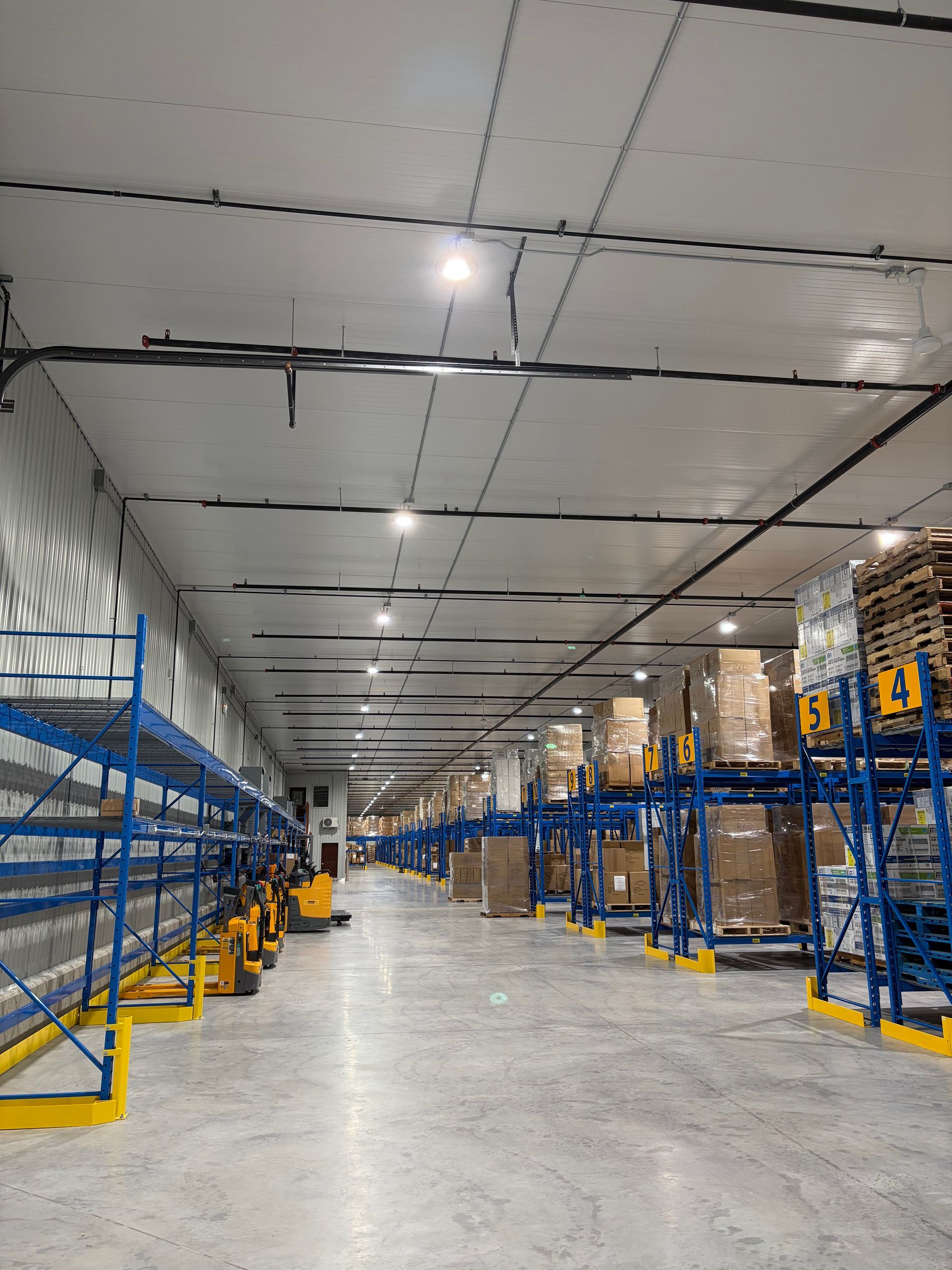Wide warehouse with blue pallet racks, stacked boxes, and bright ceiling lights