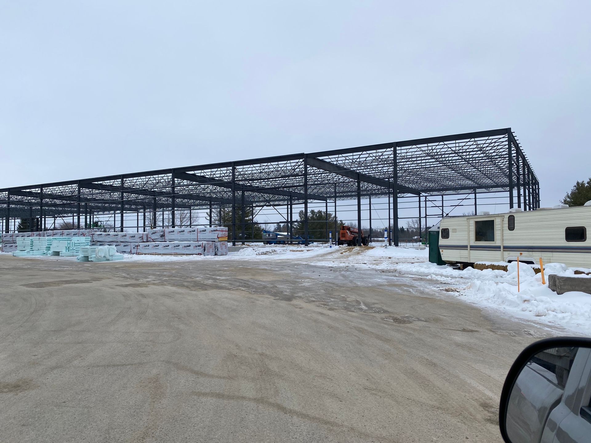 Snowy construction site with large steel building frame and parked trailers under a gray sky