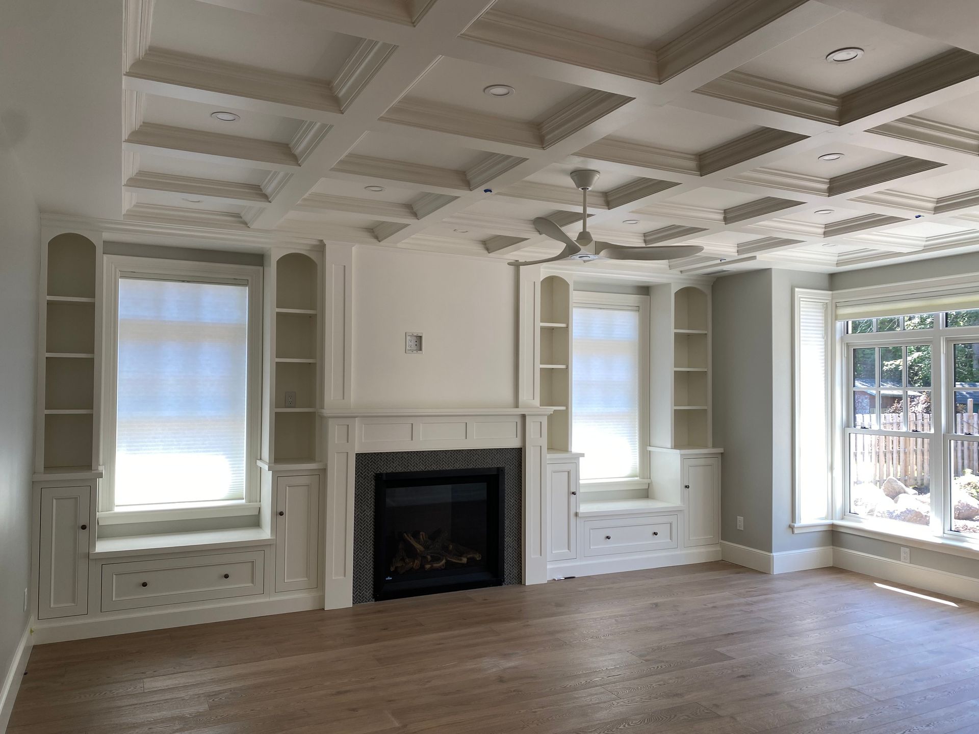 Bright empty living room with coffered ceiling, built-in shelving and storage cabinets, electric fireplace and large windows.