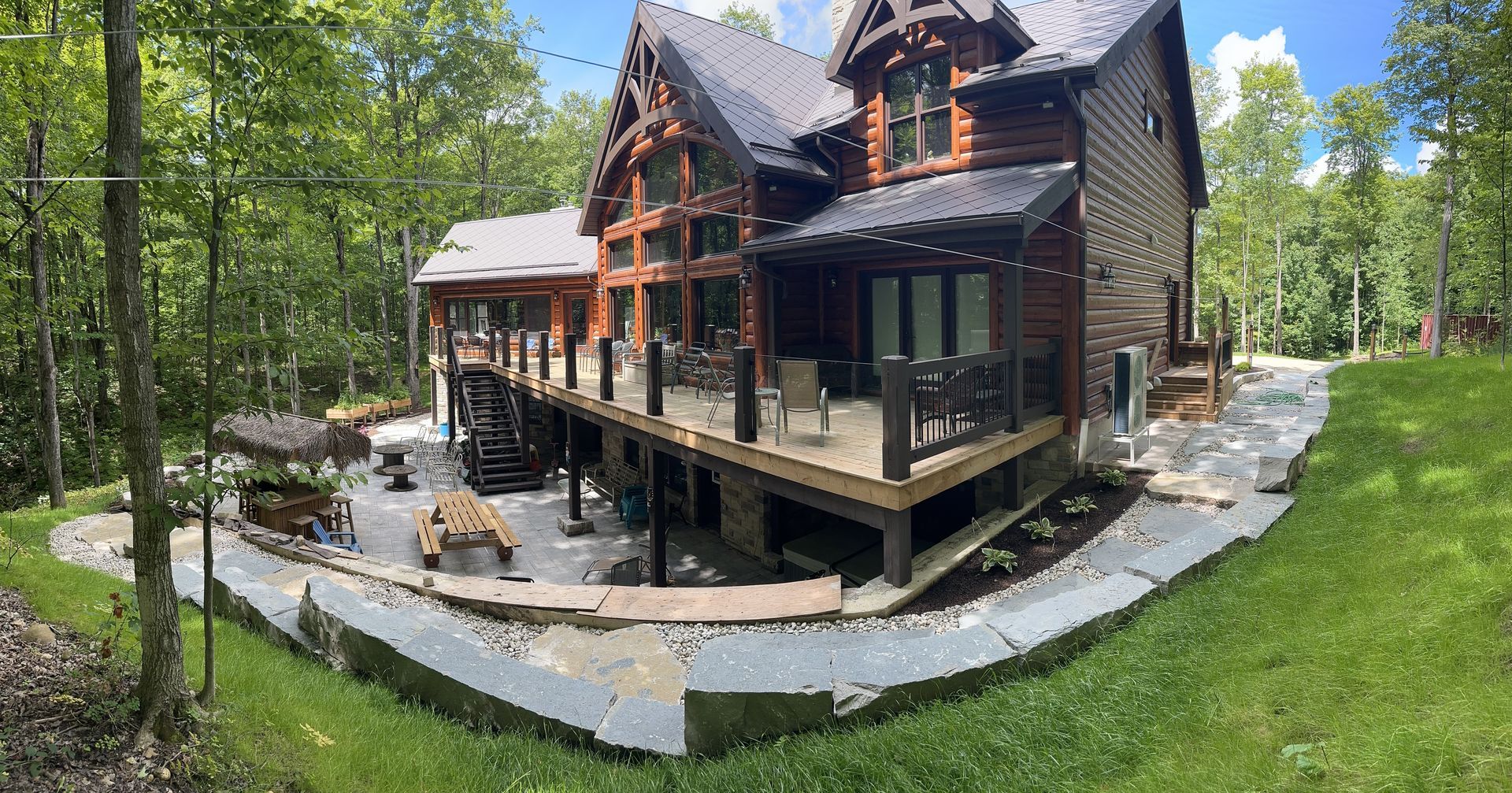 Rustic log cabin-style home with large raised porch in a wooded yard with stone patio and seating area