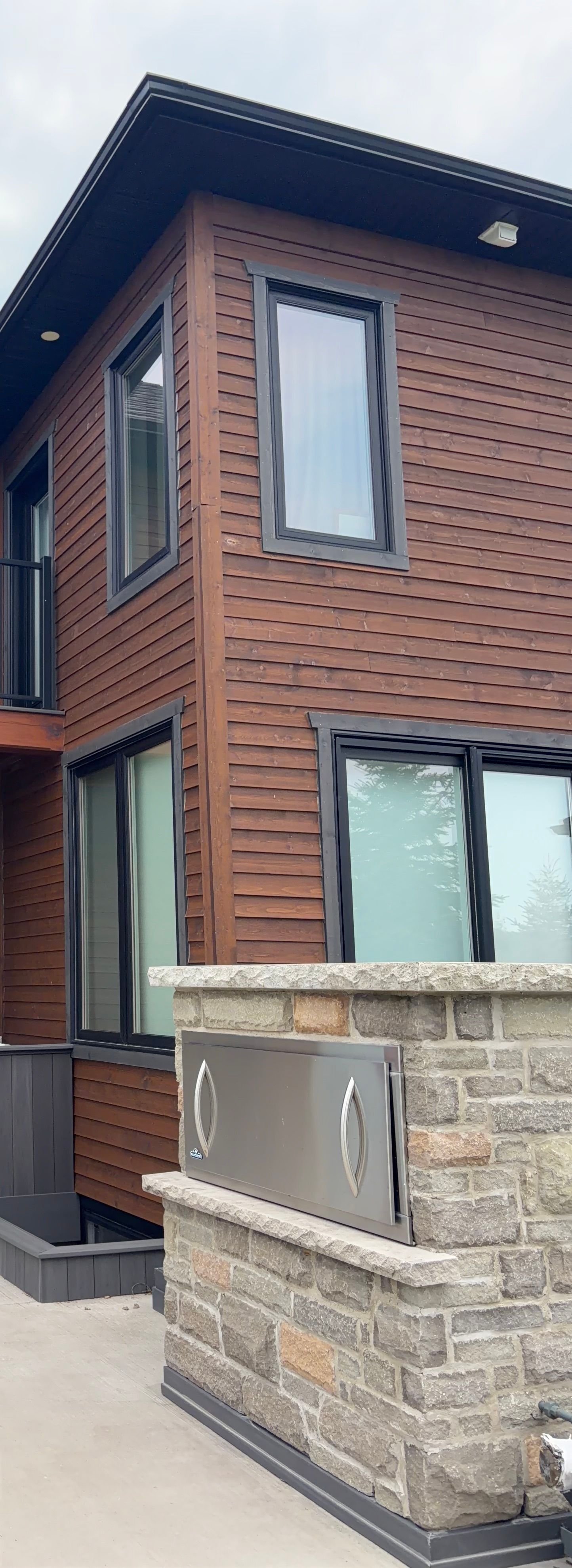 Modern house exterior with dark brown siding, large windows, and a stone walkway.