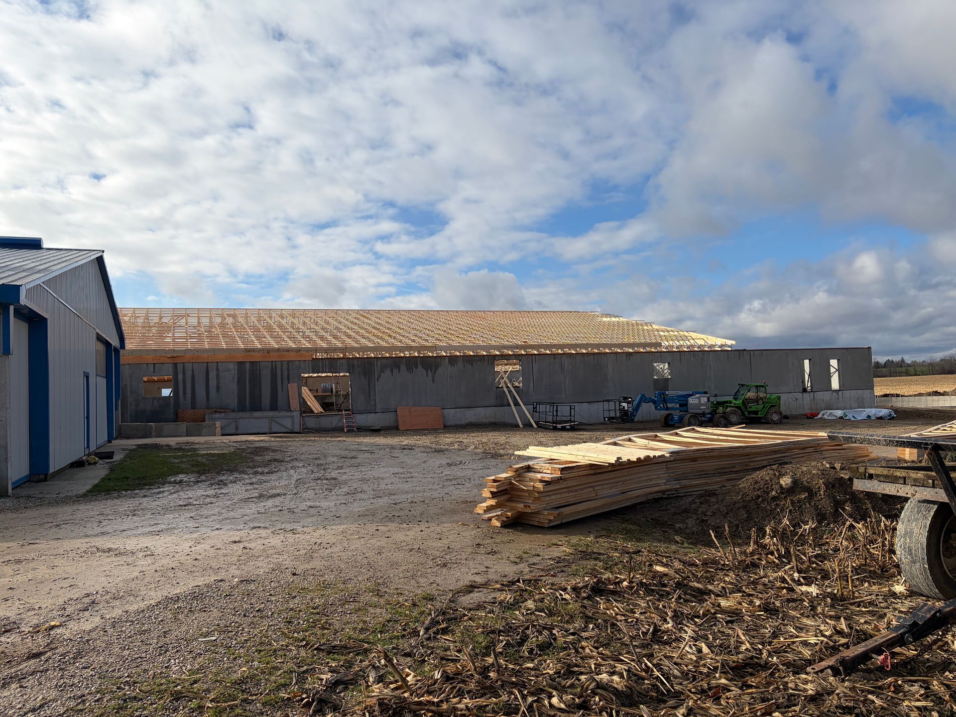 Agricultural outbuilding under construction with a partially framed building and stacked lumber under a cloudy sky