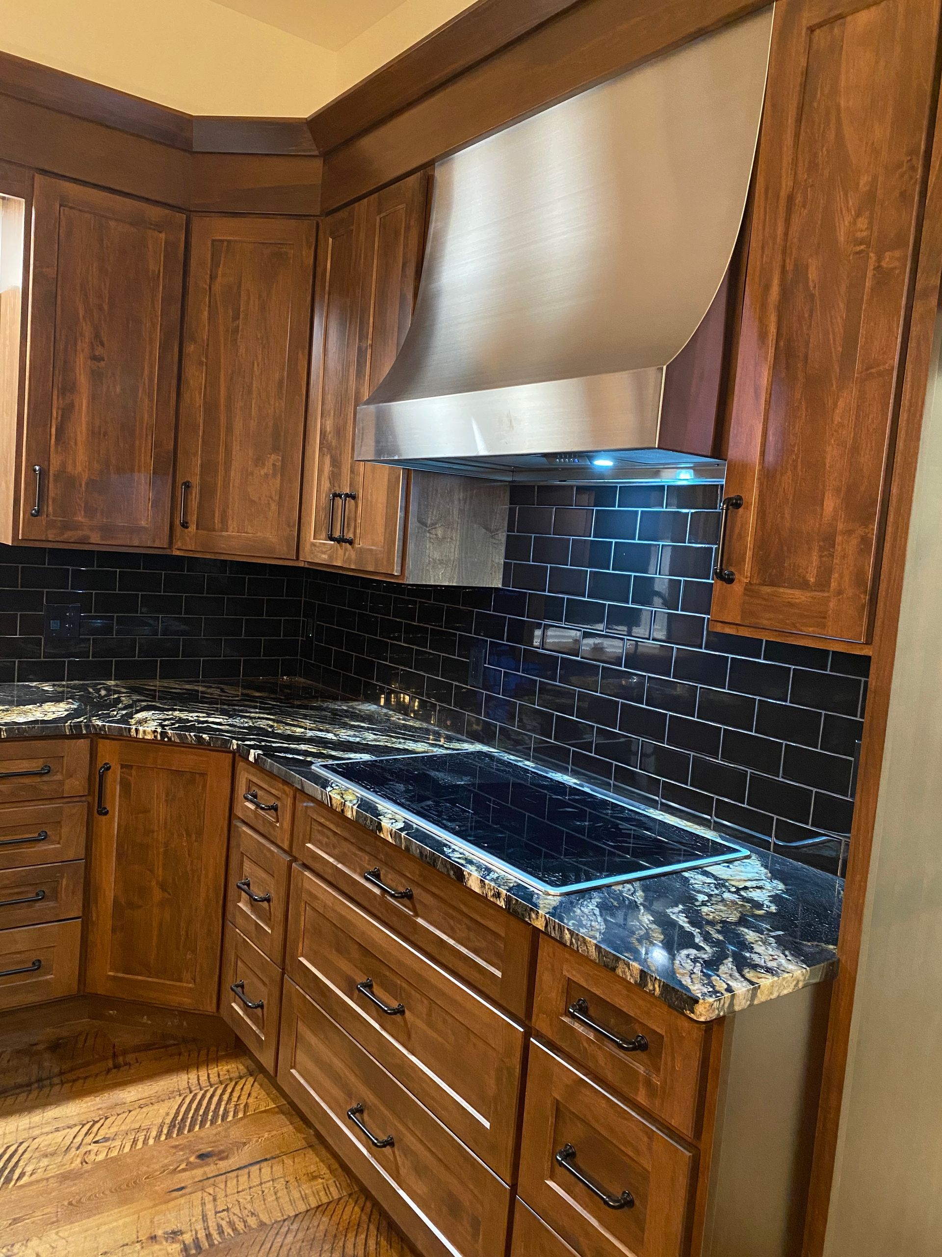 Wooden kitchen with black tile backsplash, dark marbled countertops, and stainless steel range hood.