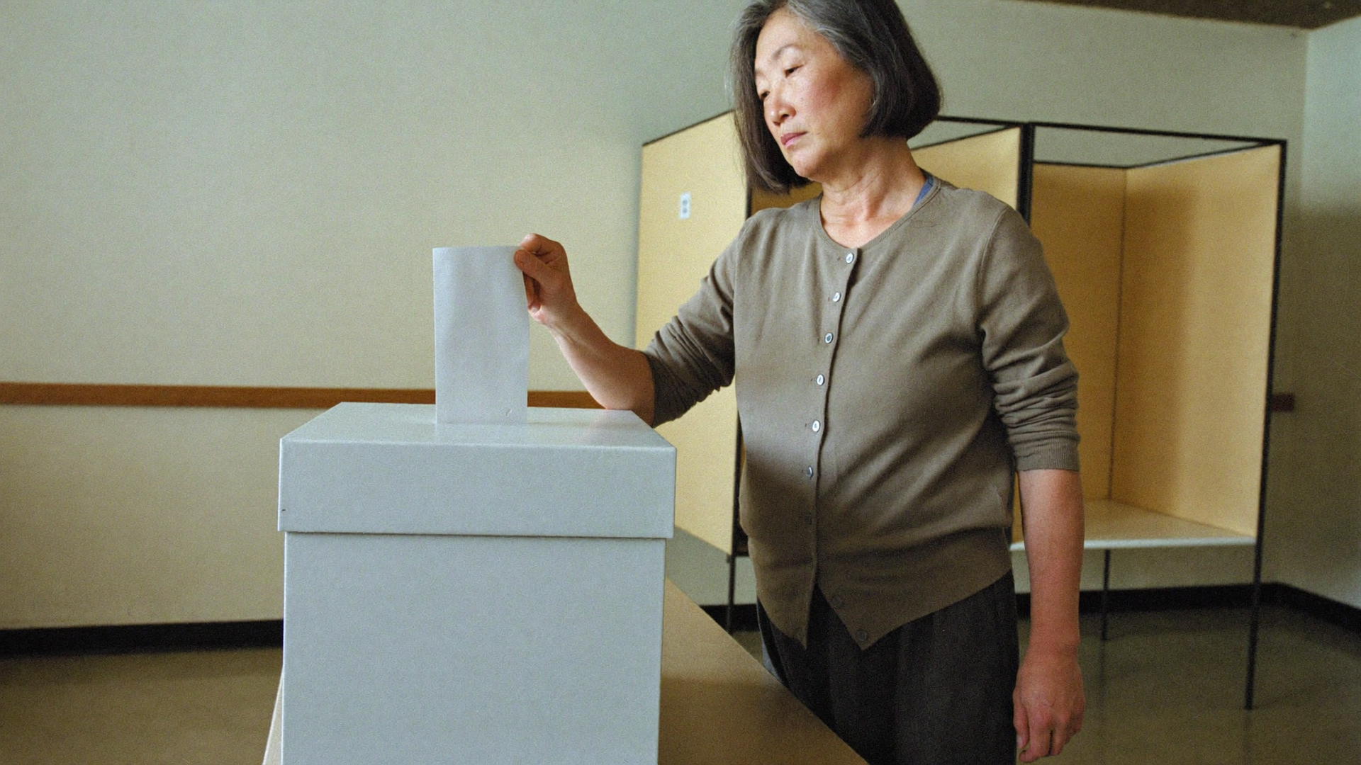Woman in a room placing a ballot into a white voting box.