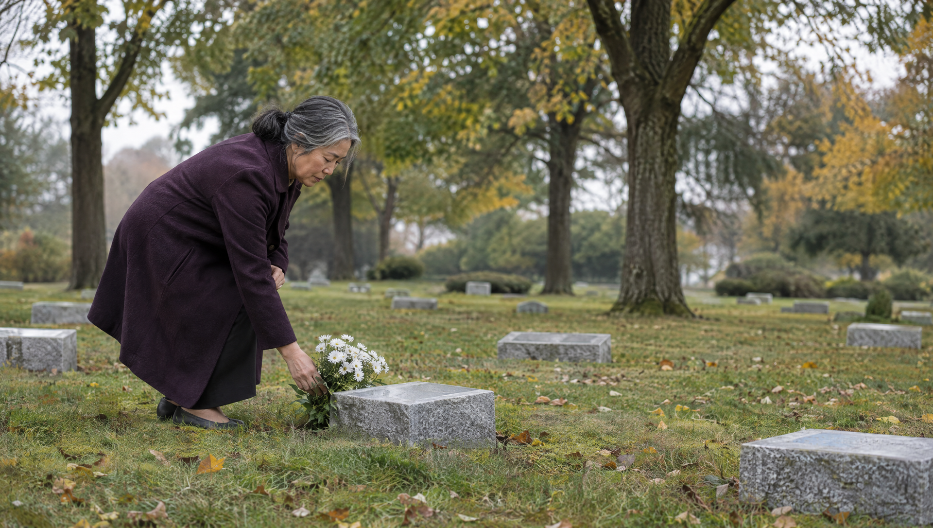 Person kneels by a grave placing flowers in a cemetery under overcast trees