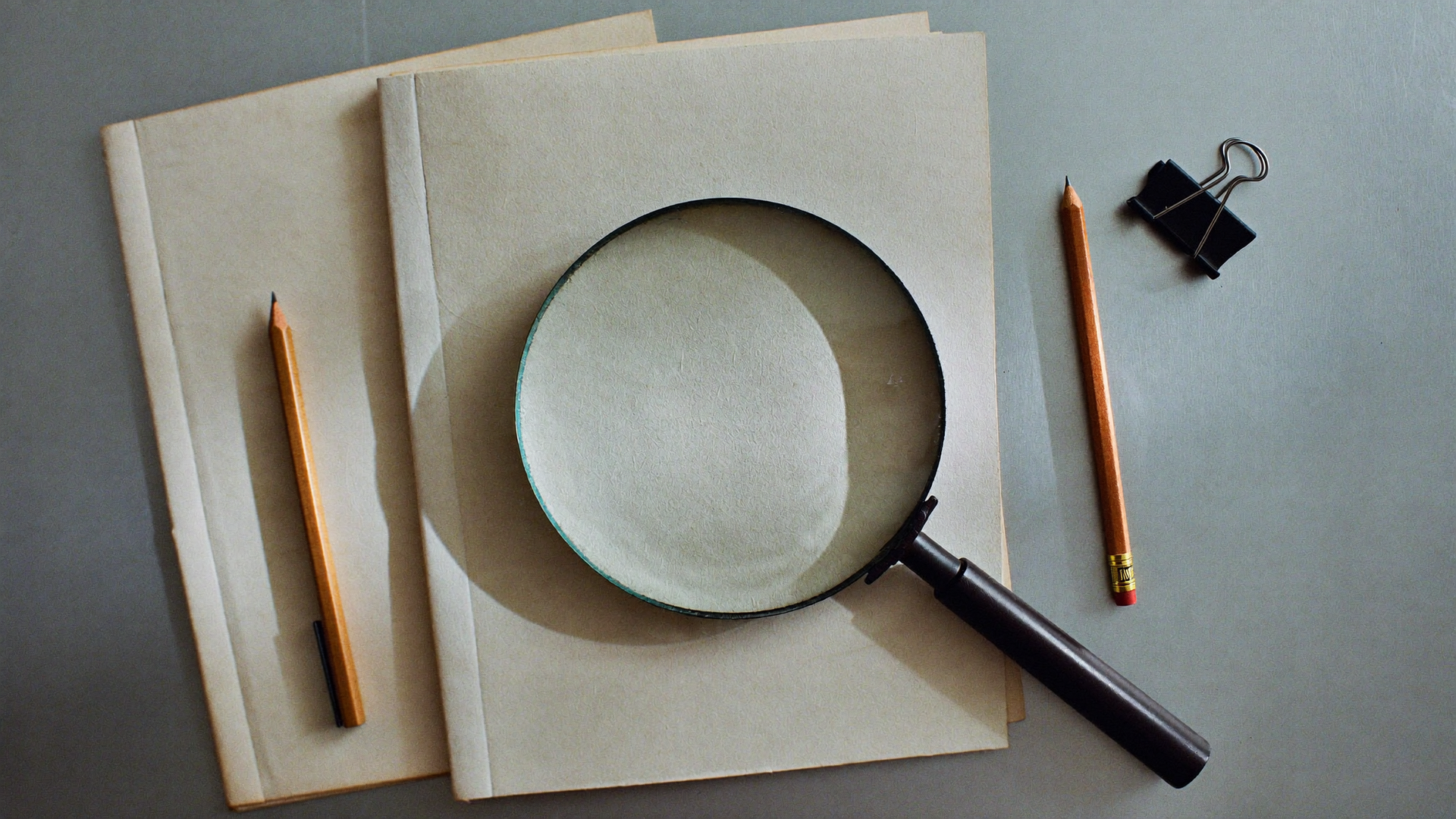 Notebook, magnifying glass, pencils, and paper clips on a gray desk.
