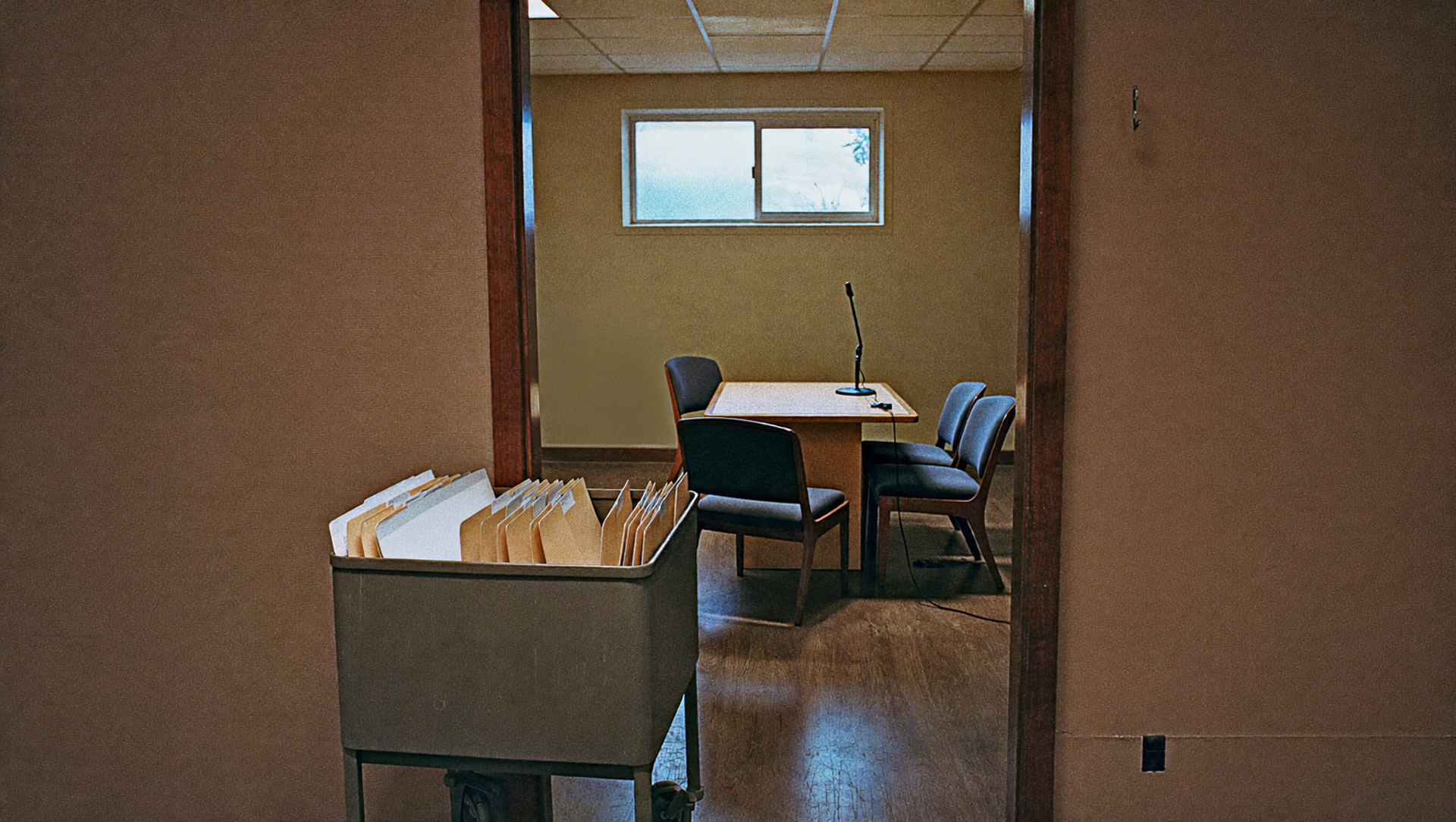 Small office with desks, chairs, and a filing cabinet in a dim hallway view