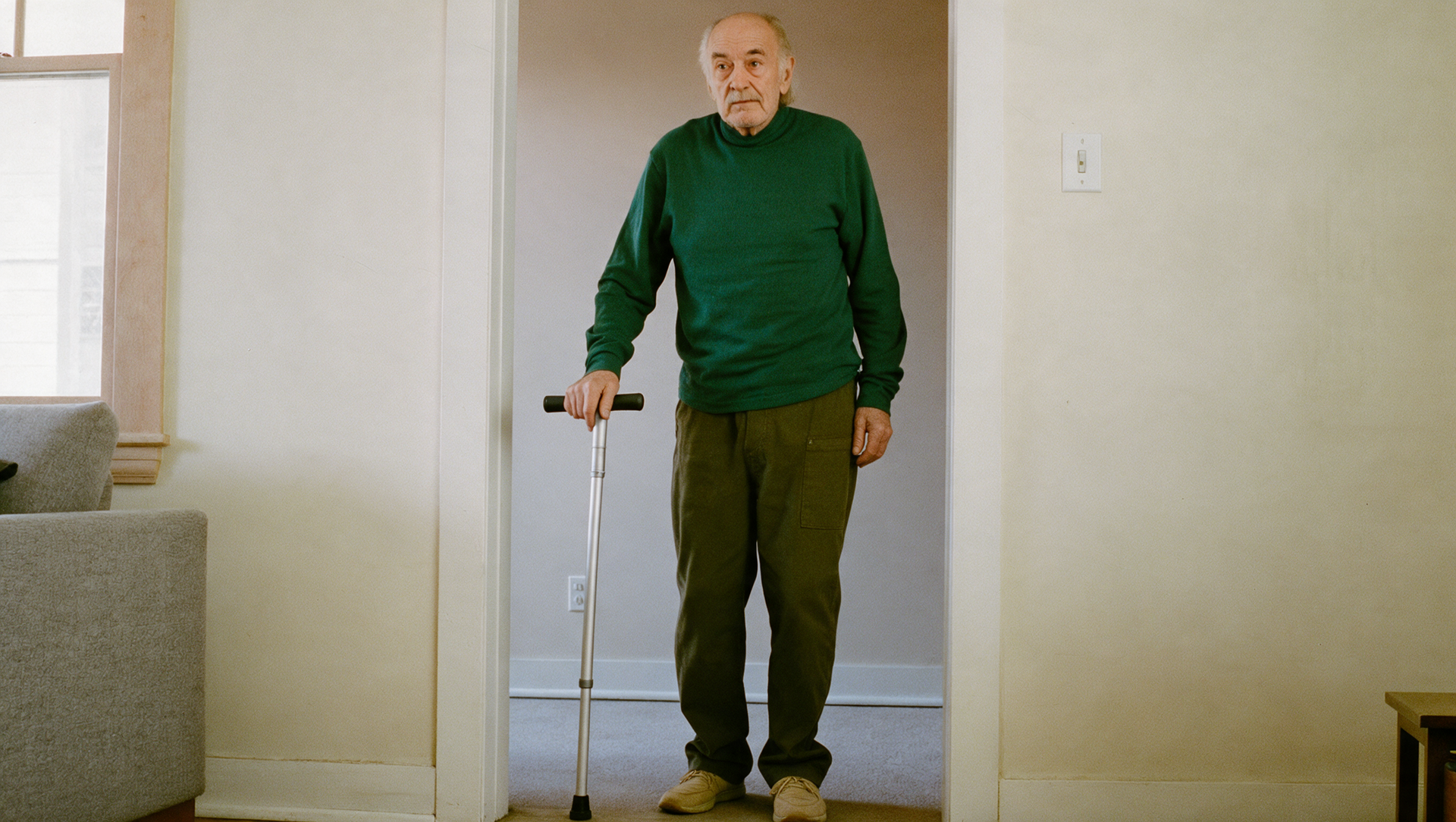 Man standing in a doorway holding a cane, wearing a green sweater and brown pants.