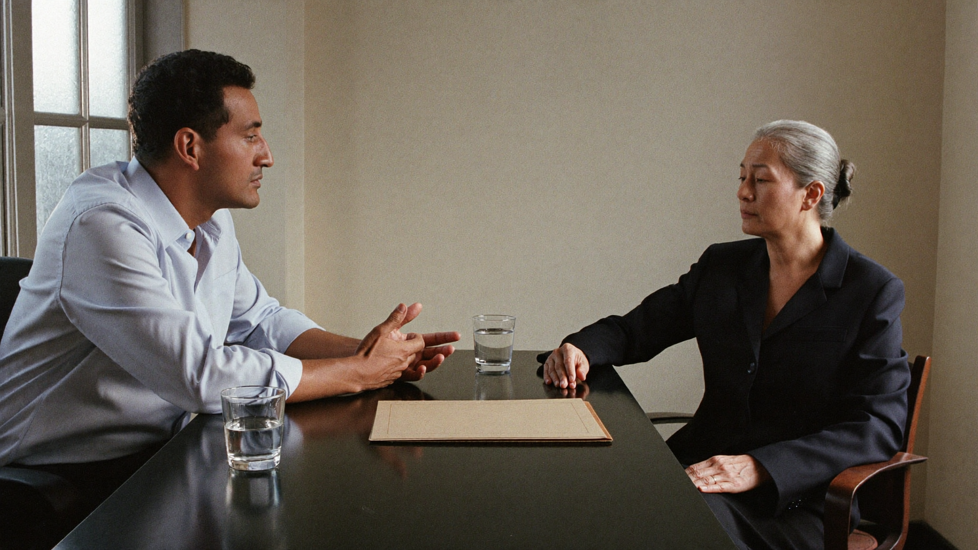 Two people in a business meeting across a table, talking with a notepad and glass of water between them