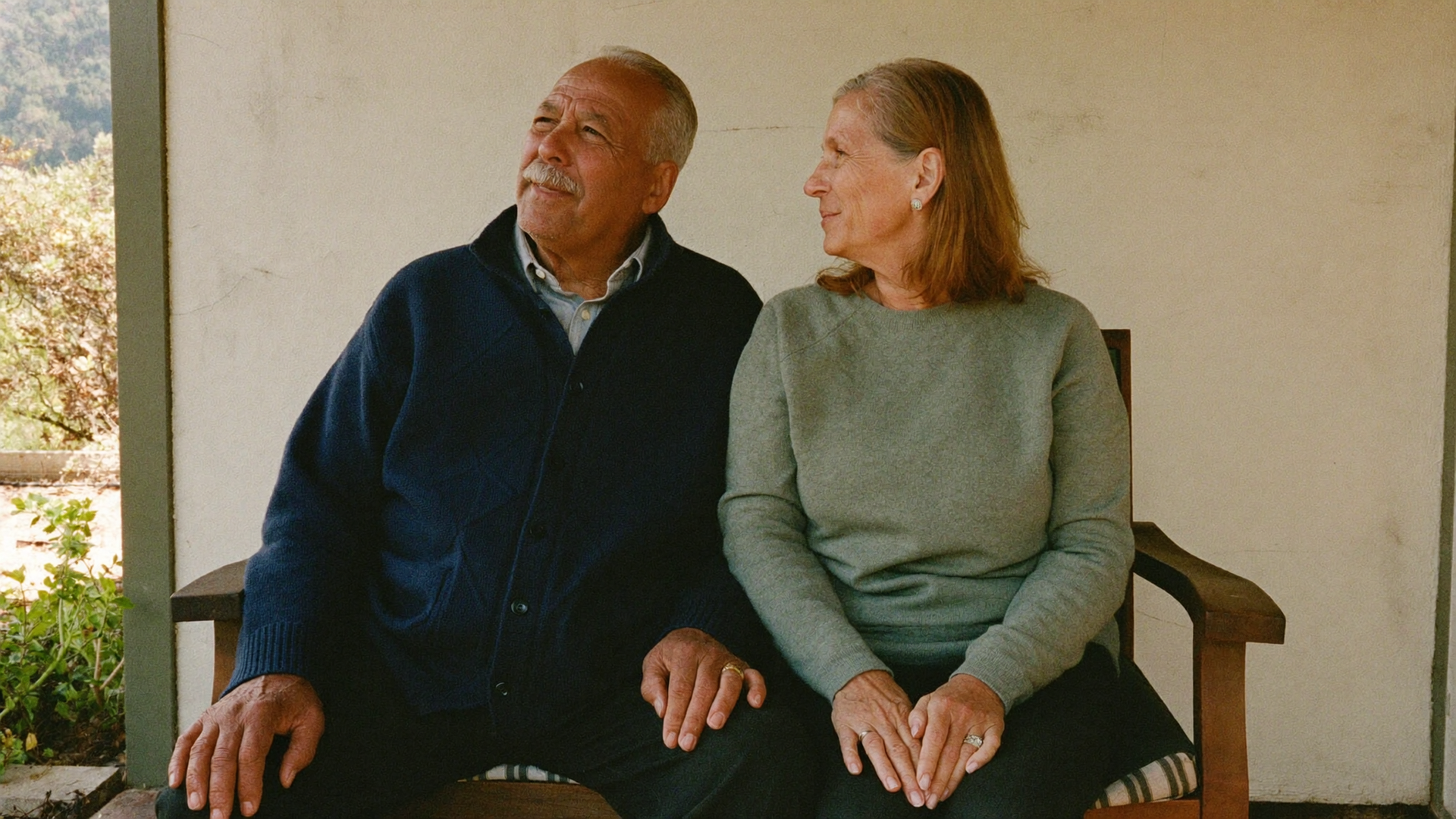 Two people sitting on a wooden porch bench, looking off to the left, with a beige wall behind them.
