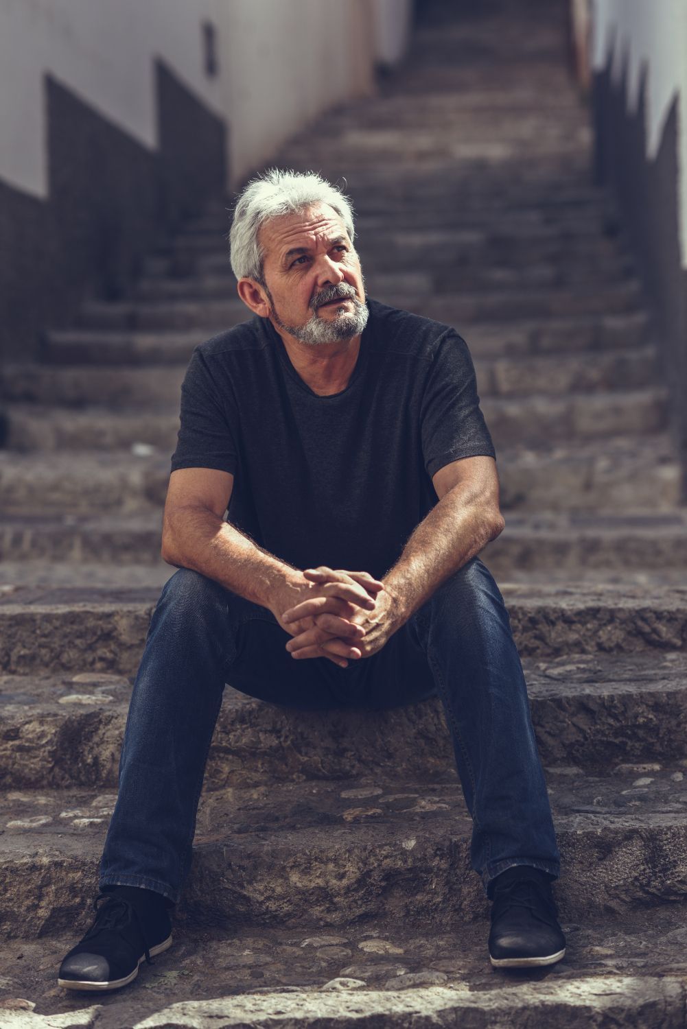 Man seated on outdoor steps, wearing dark clothes, looking off to the side with hands clasped.