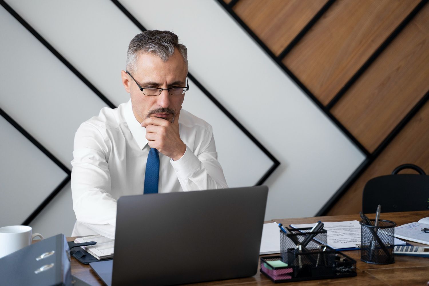 Man in white shirt and blue tie working at a laptop, looking thoughtful at a desk with office items