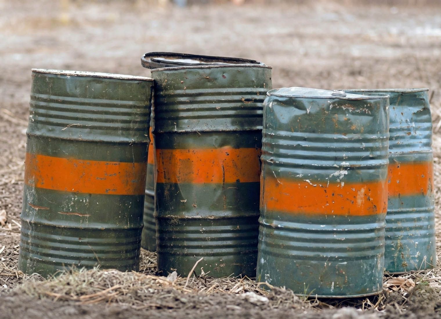 Three weathered metal barrels with orange bands standing outdoors on dirt