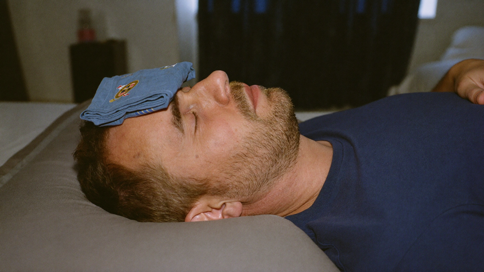 Man lying on a couch with a blue cloth over his forehead and eyes, resting indoors.