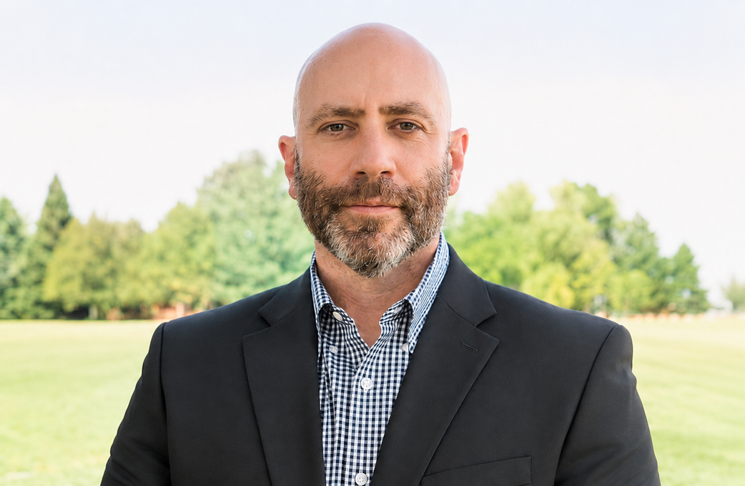 Portrait of a bald man in a blue blazer and white shirt outdoors with blurred trees behind him
