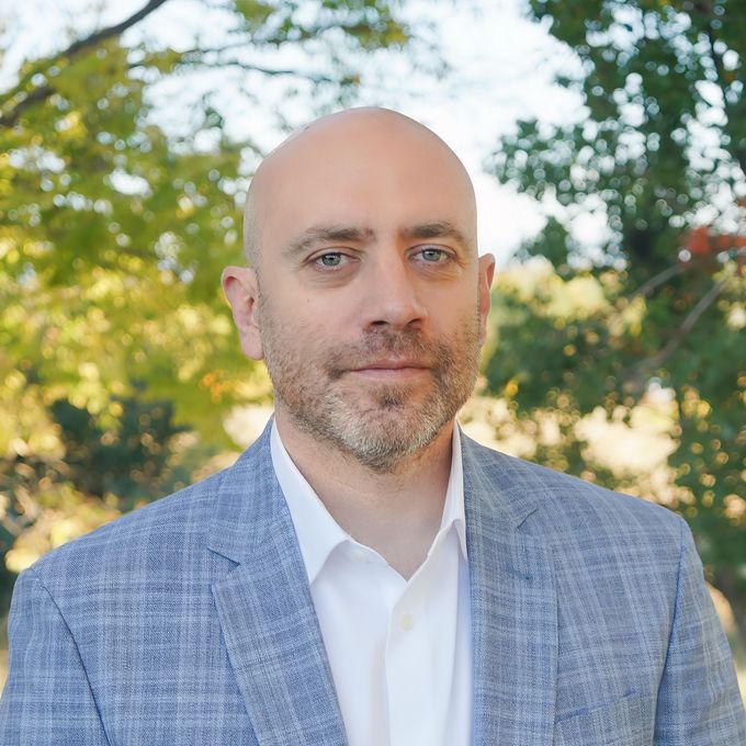 Portrait of a bald man in a blue blazer and white shirt outdoors with blurred trees behind him