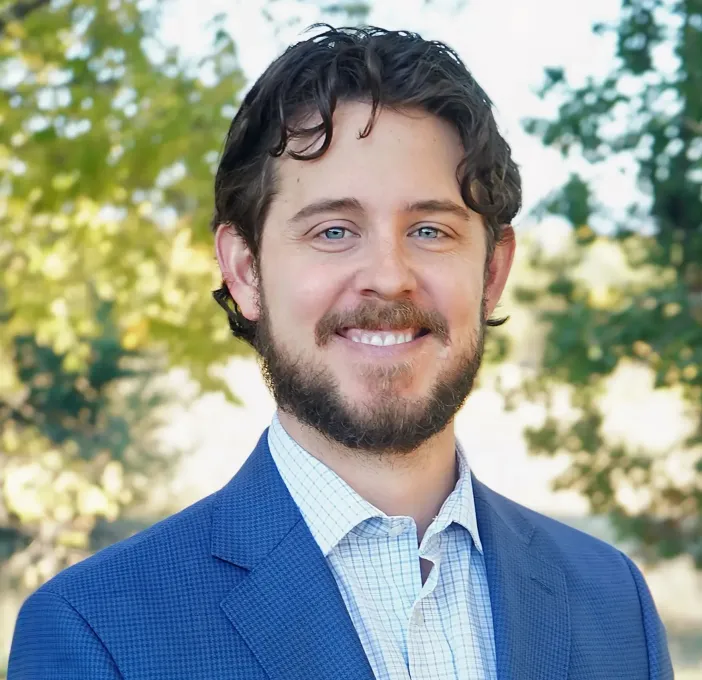 Smiling man in a blue blazer outdoors with blurred trees in the background