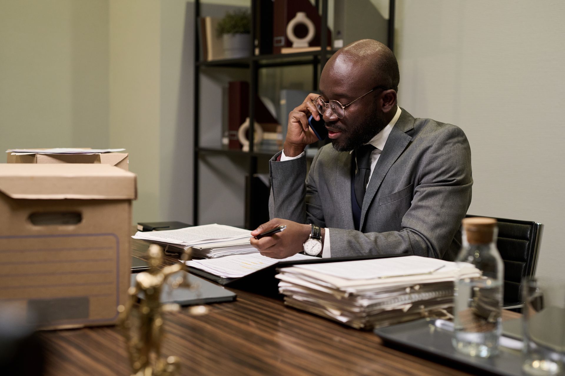 Man in a suit talking on a phone at a desk with stacks of papers and a printer in an office