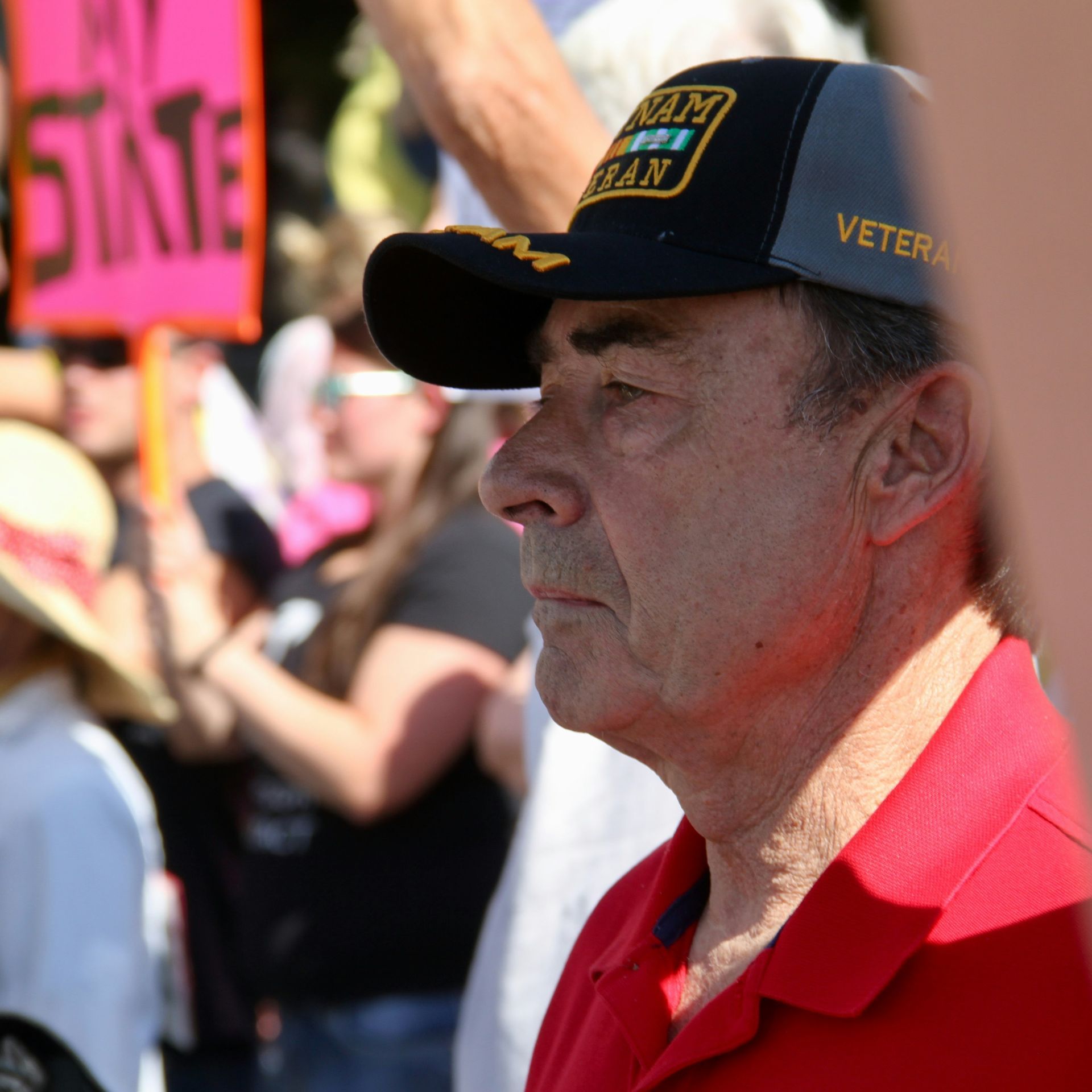 Man in red polo and navy cap at an outdoor crowd, in profile looking left