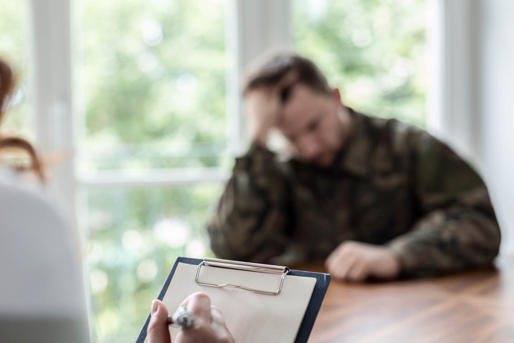 Clipboard with notes in foreground; blurred person in camouflage sits at a table in a bright room.