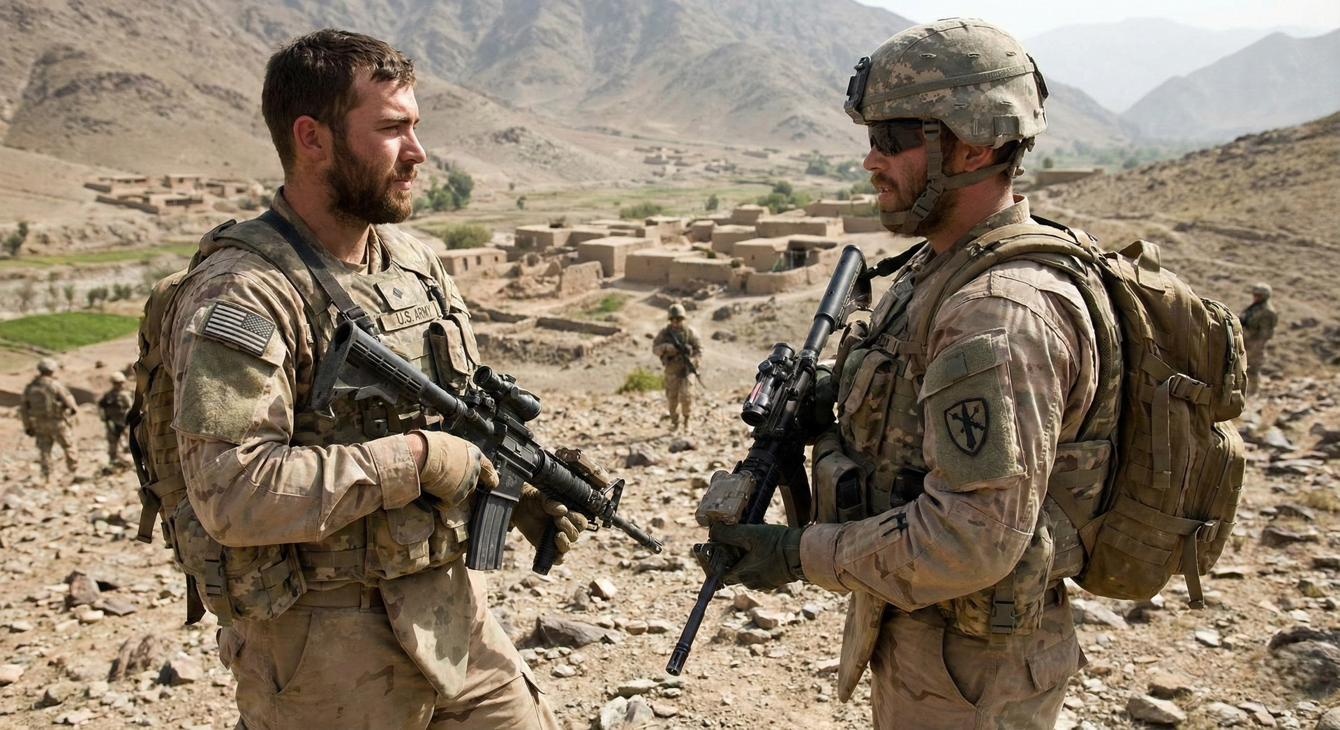 Two soldiers in camouflage gear talk in a rocky desert valley with mountains and tents behind them