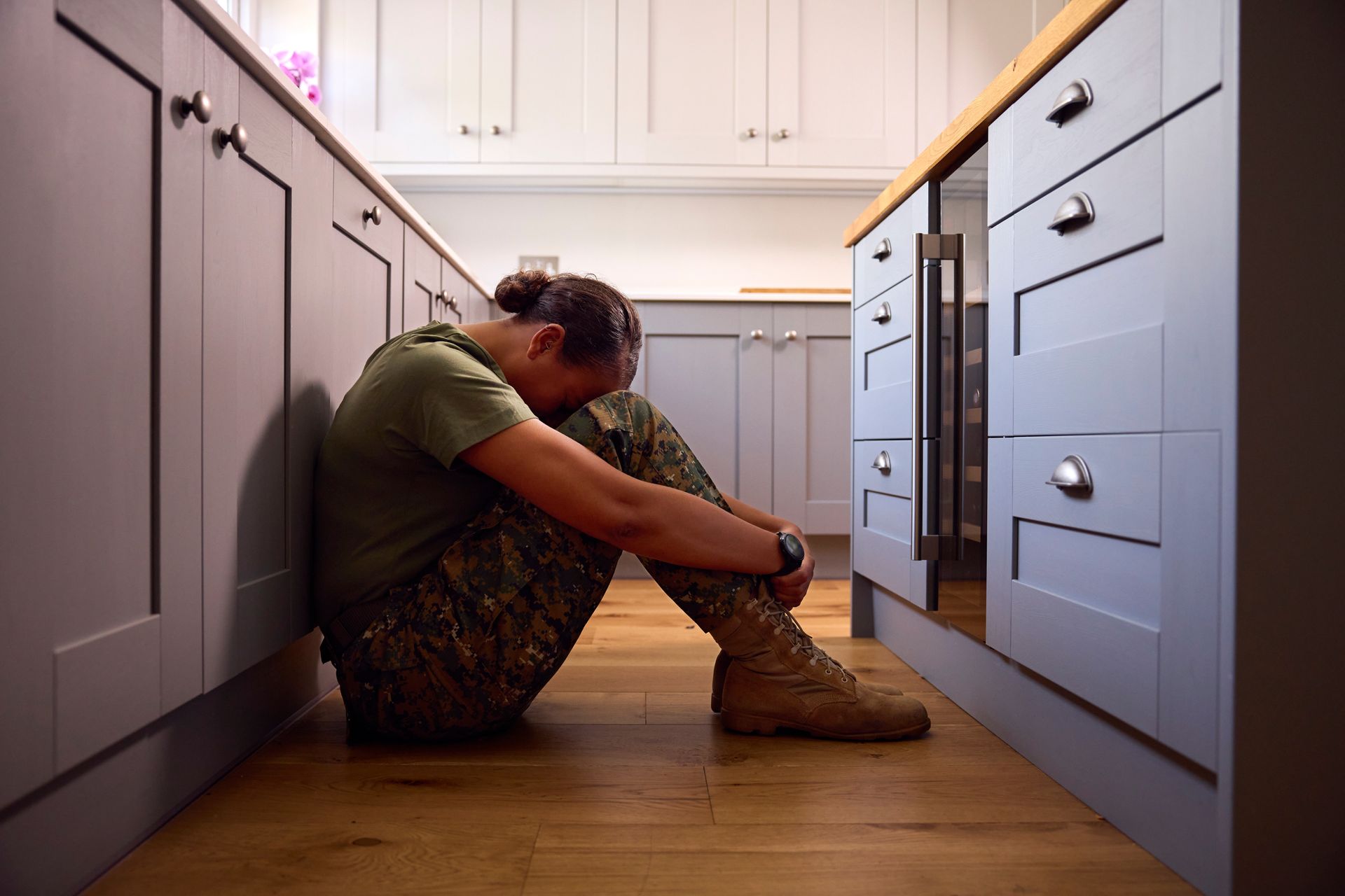 Person sitting on a kitchen floor with head down, arms wrapped around knees, beside white cabinets.