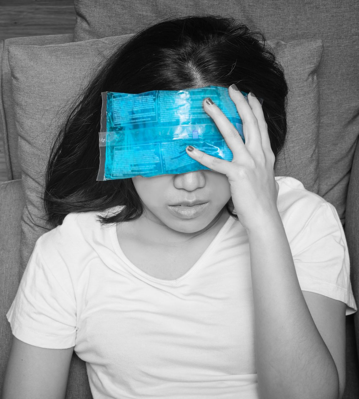 Person lying on a bed, holding a blue ice pack over their eyes.