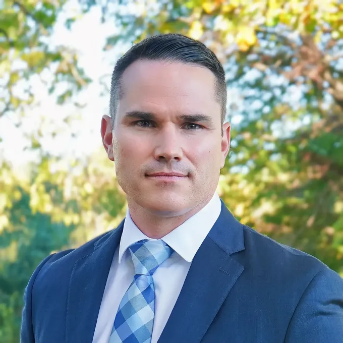 Professional man in a blue suit and tie outdoors, standing before a blurred tree-lined background