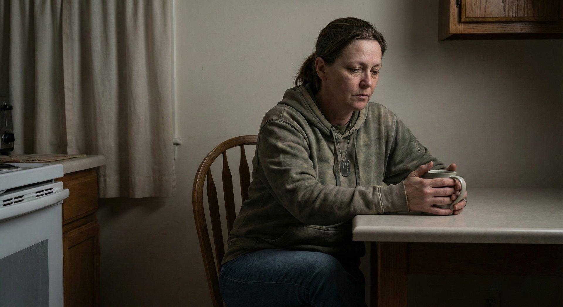Woman sitting at a kitchen table, hands clasped, looking pensive beside a window and stove