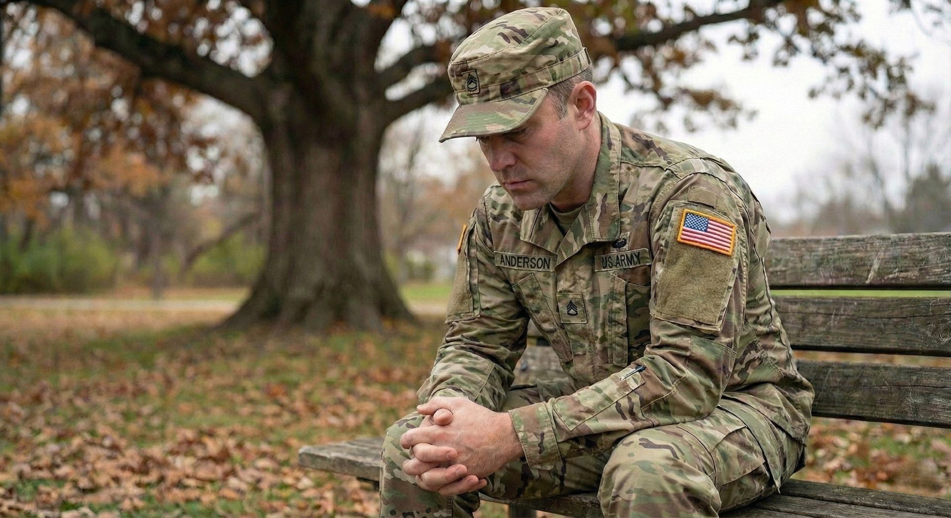 Soldier in camouflage uniform sitting on a bench outdoors, head bowed in a quiet, reflective pose.