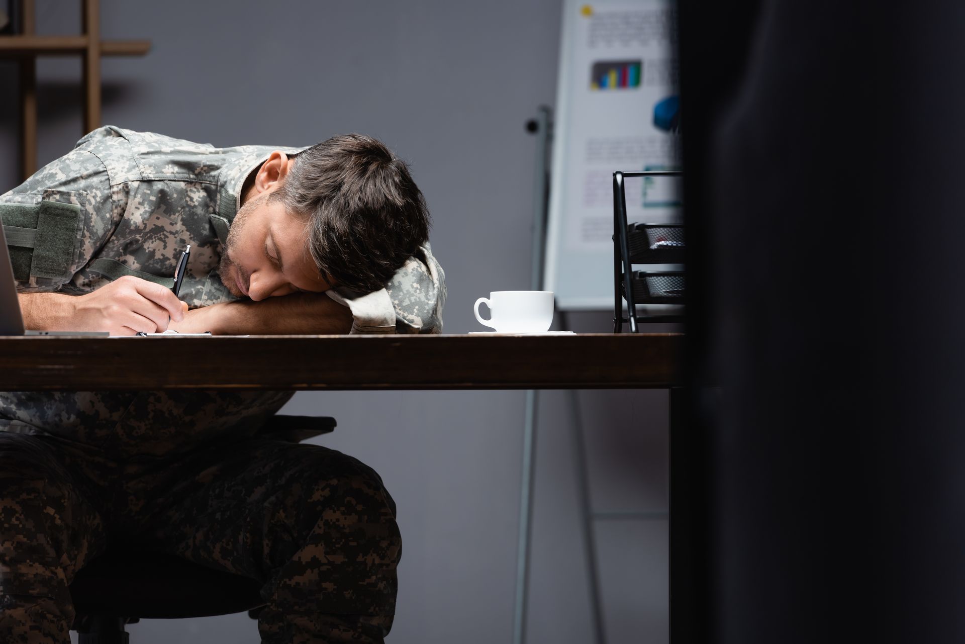 Person asleep at a desk in a dim office, head resting on folded arms beside a coffee cup.