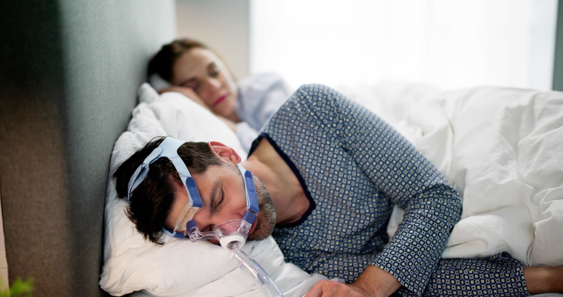 Patient wearing a CPAP mask lying in bed, with a person resting behind them in a softly lit room