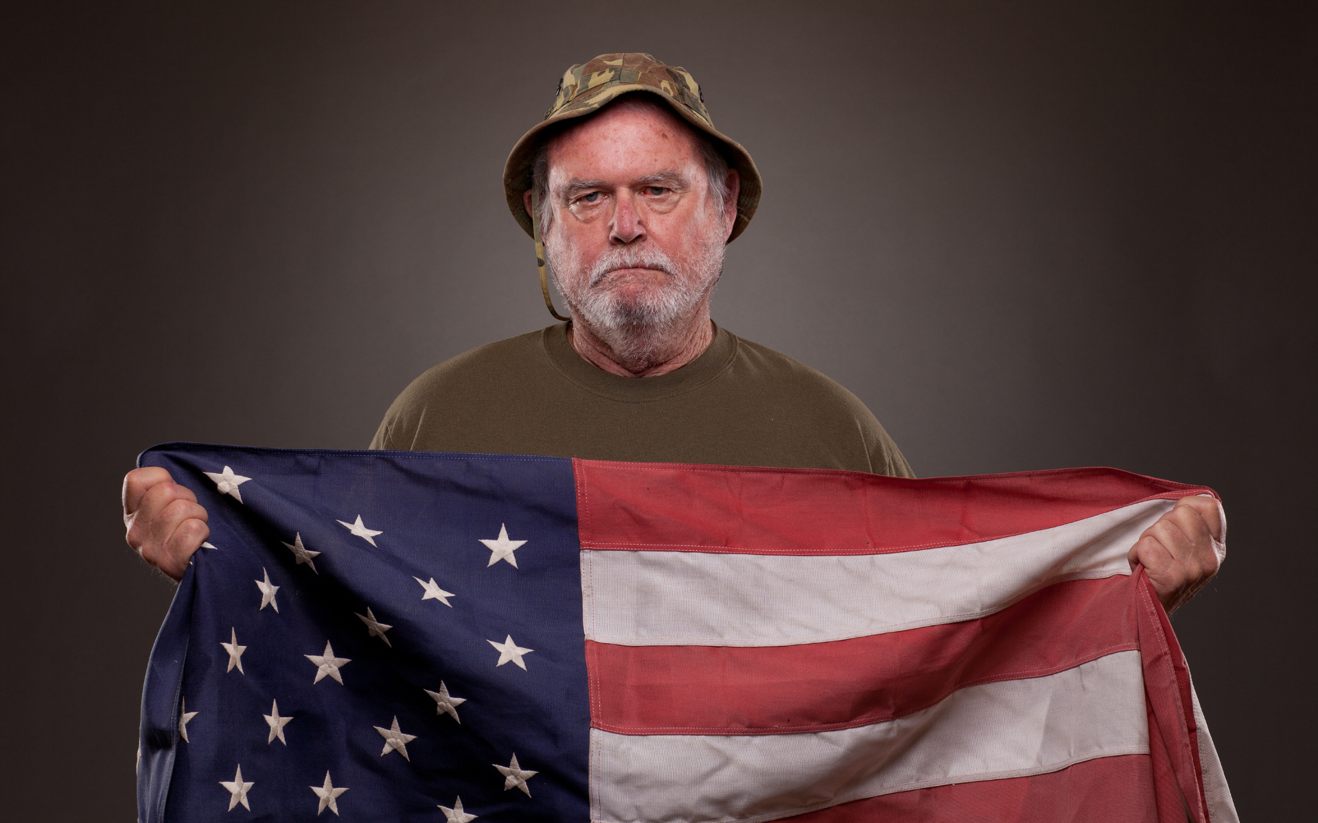 Man in a camo hat holding a U.S. flag against a plain dark background