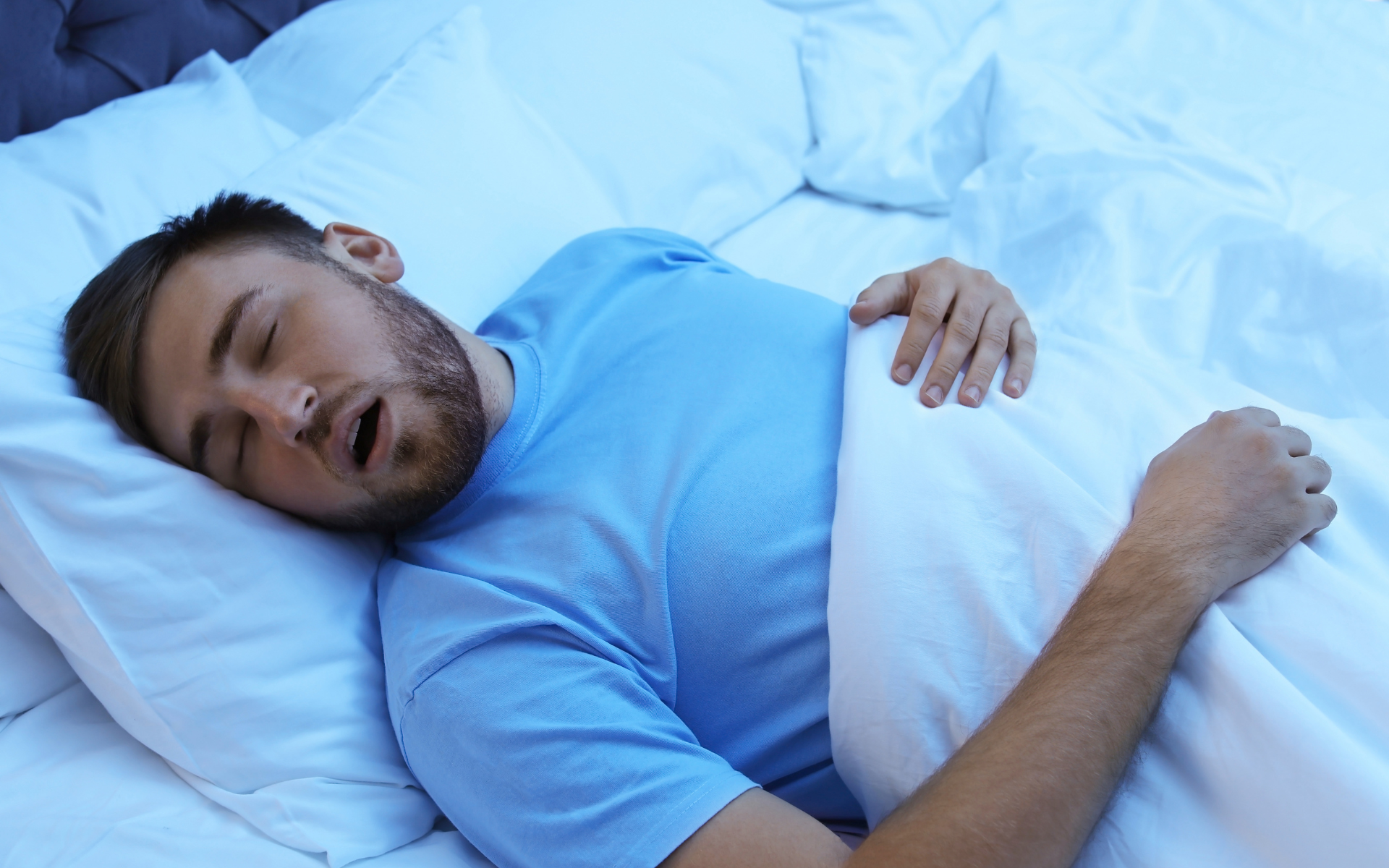 Sleeping man in a light blue shirt lying on white bedding with one hand on his chest