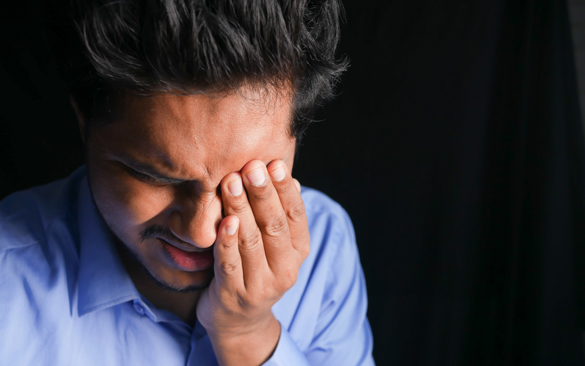 Man in a blue shirt holding his face, eyes closed, looking distressed against a dark background
